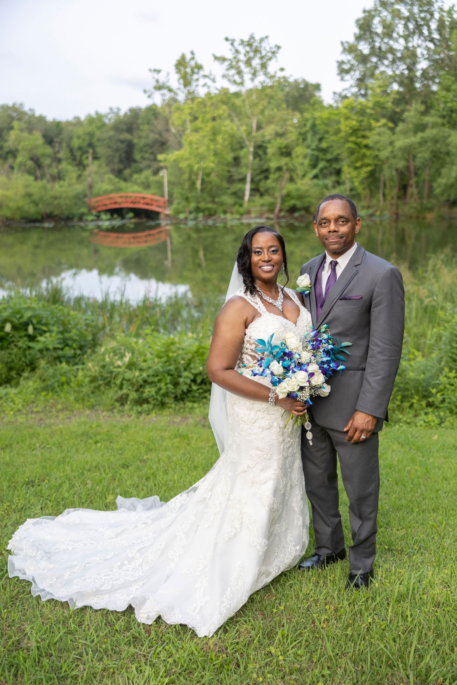 A couple in wedding attire poses together on a grassy lawn near a pond with trees in the background.