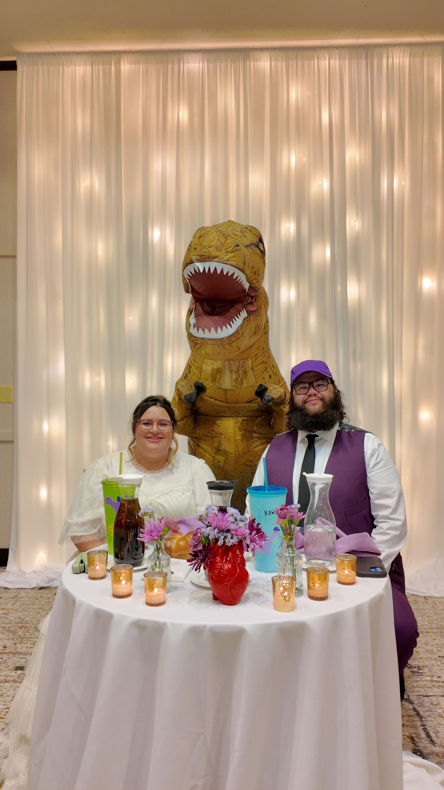 A wedding party at a reception table features a unique dinosaur costume and colorful attire against a white backdrop with lights.
