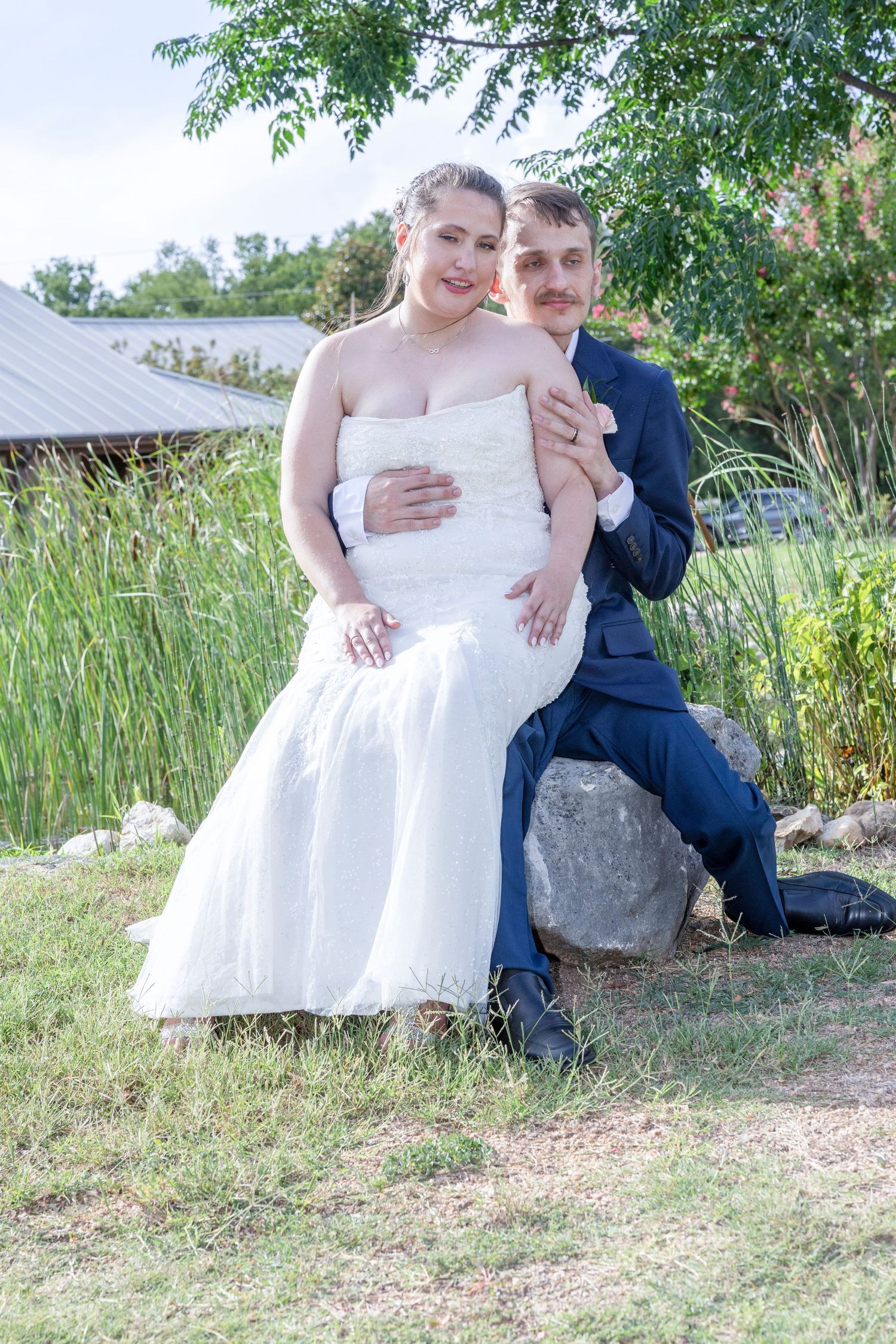 A couple in wedding attire sits together on a stone wall surrounded by lush greenery in an outdoor setting.
