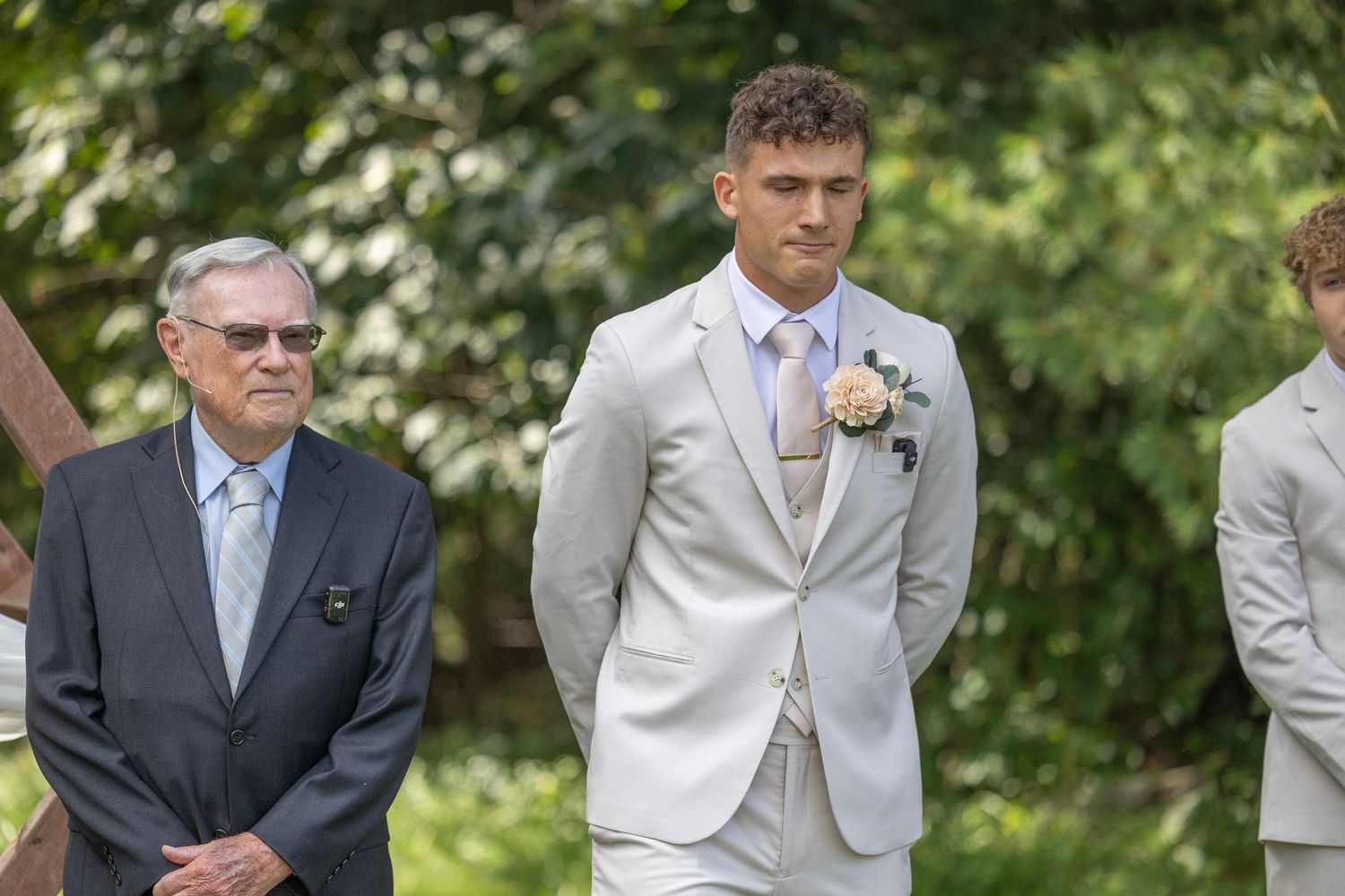A group of groomsmen in light and dark suits stand together outdoors in a garden setting.