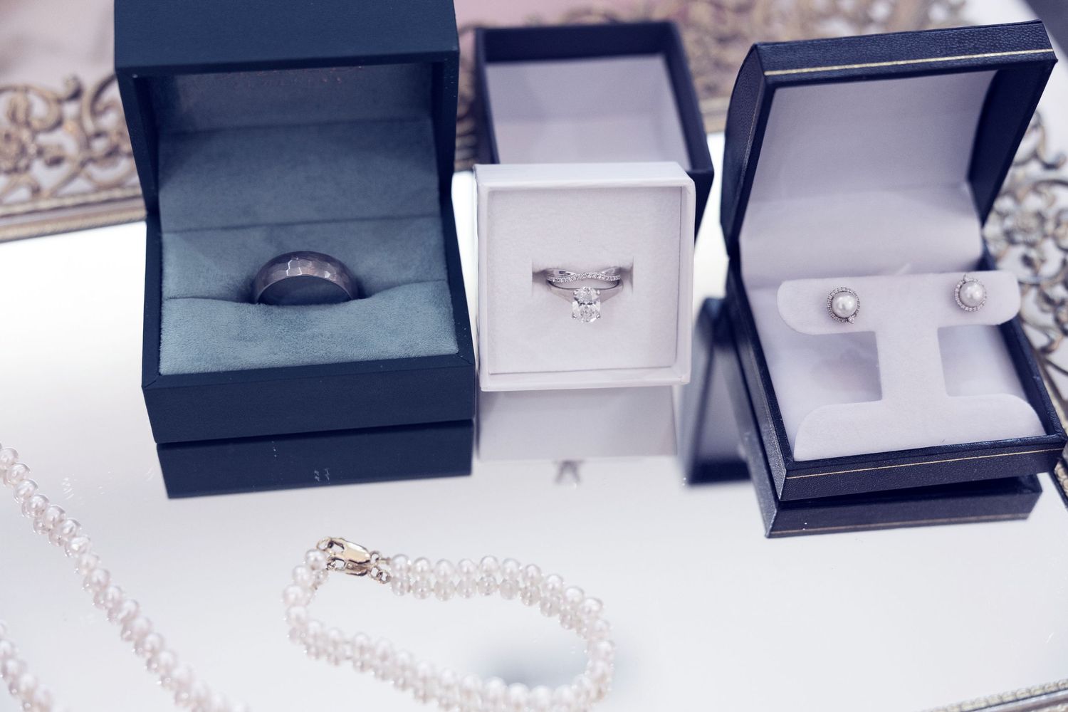 Three elegant jewelry boxes displaying diamond rings and pearl earrings, with a delicate pearl bracelet on a mirrored surface.