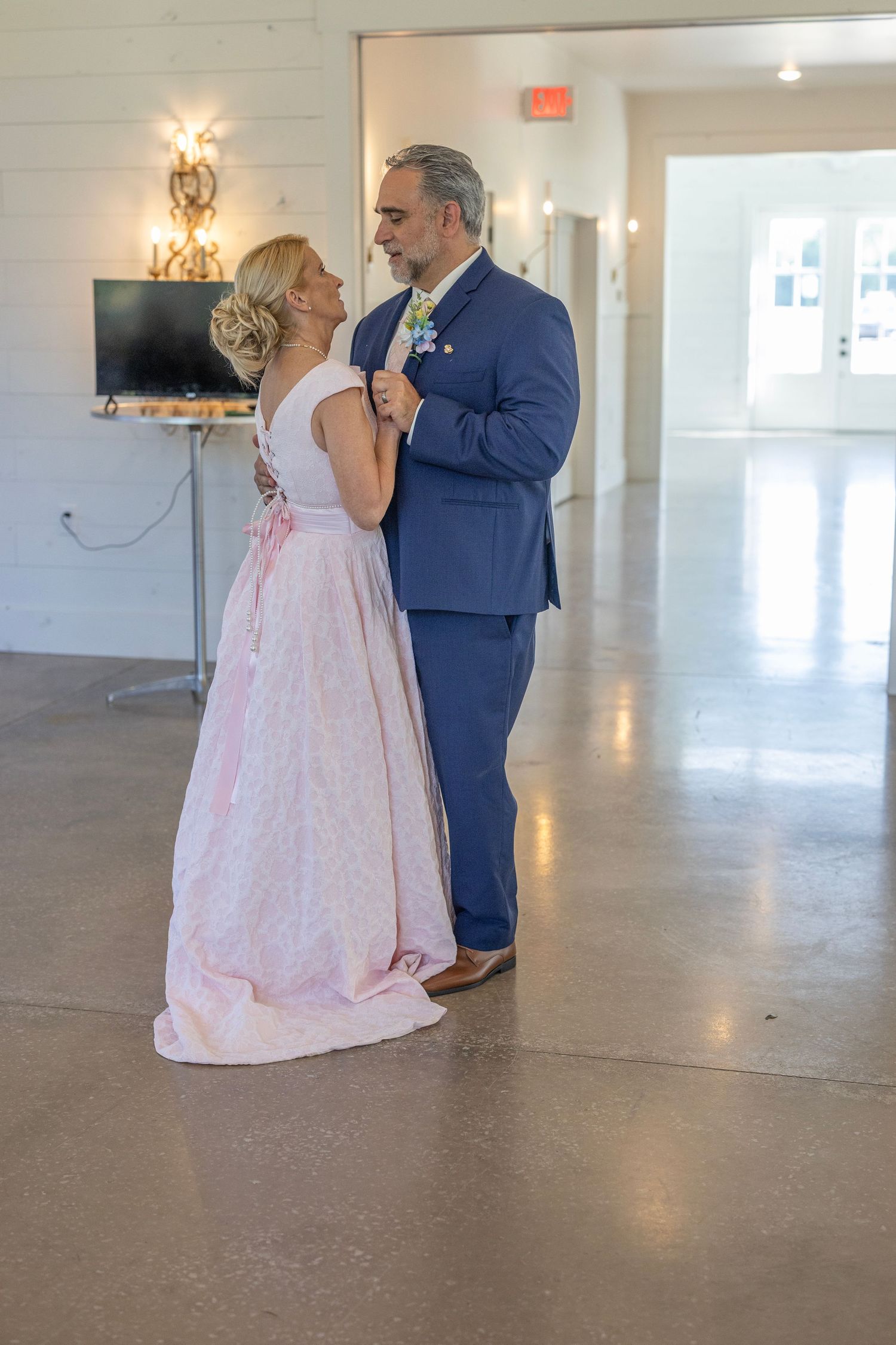 Couple sharing first dance at wedding, woman in pink gown and man in navy suit, elegant white venue.
