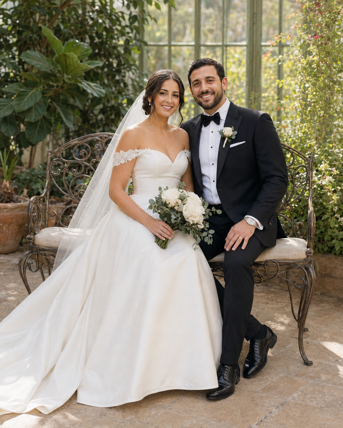 Bride and groom sitting together in a garden, bride in off-shoulder white gown holding bouquet, groom in black tuxedo.