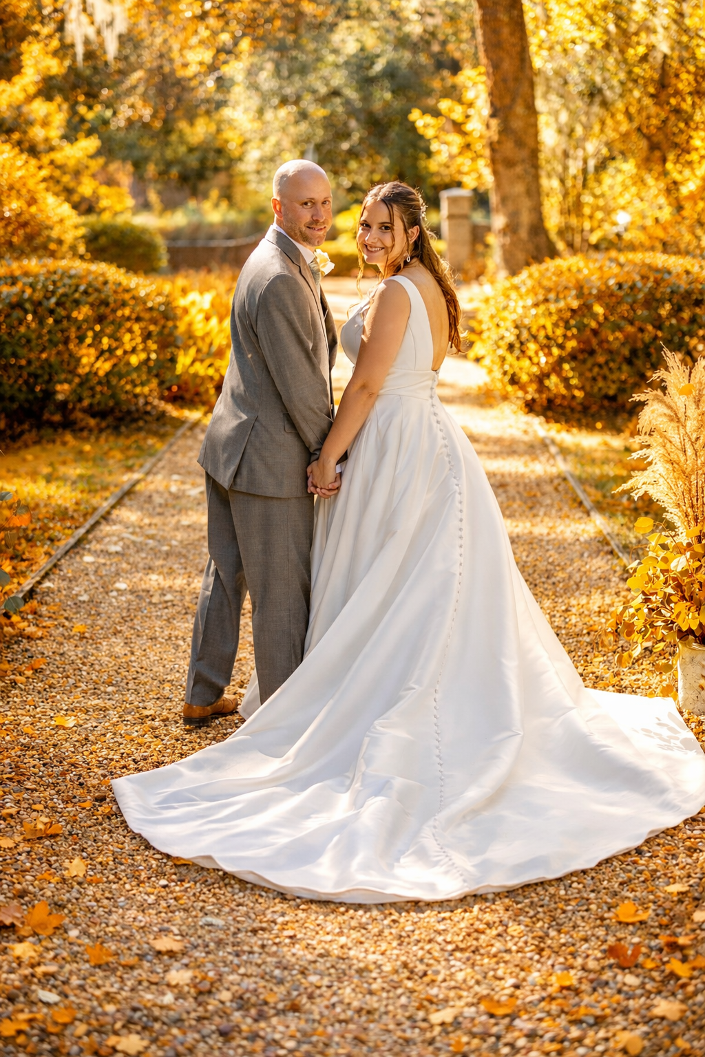 Bride and groom holding hands on a garden path surrounded by golden autumn foliage in an elegant outdoor wedding portrait.