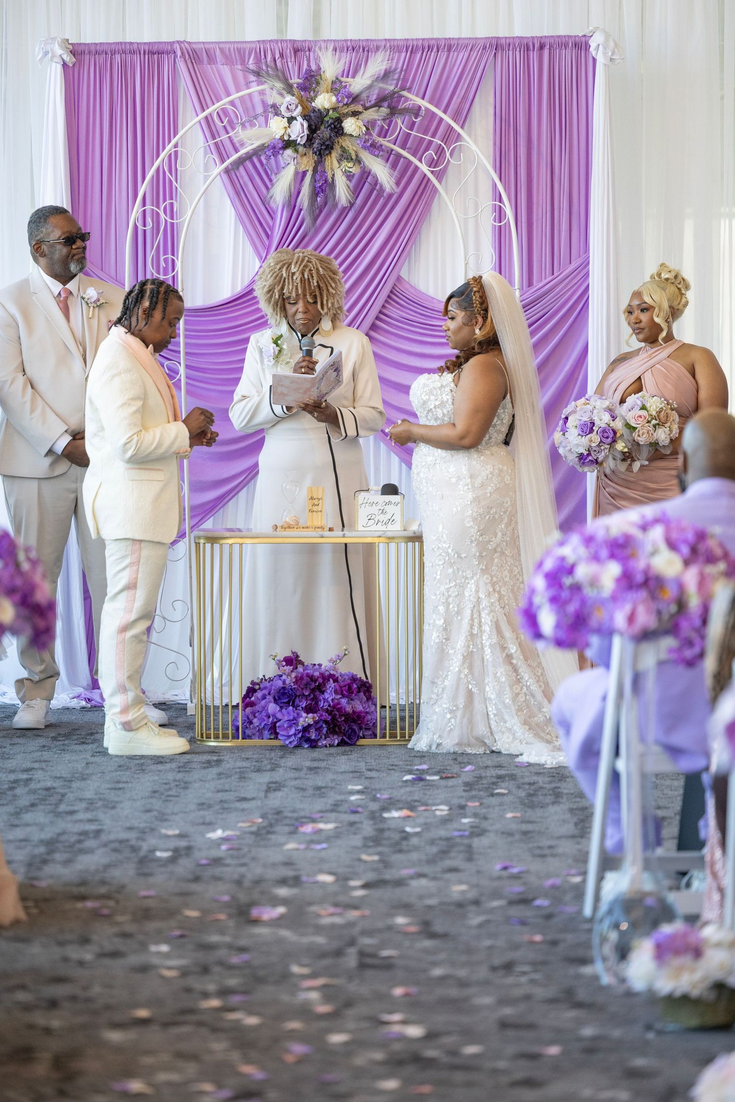 Elegant wedding ceremony with bride and groom at purple-draped floral arch altar, bridesmaid holding bouquet nearby.