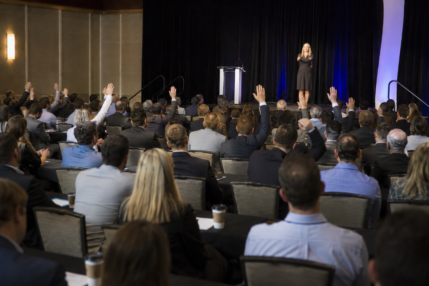 Speaker on stage engages audience with raised hands at a packed corporate conference session.
