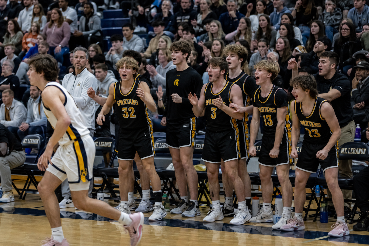 High school basketball players in black and gold uniforms celebrate on the sideline during an intense gym game.