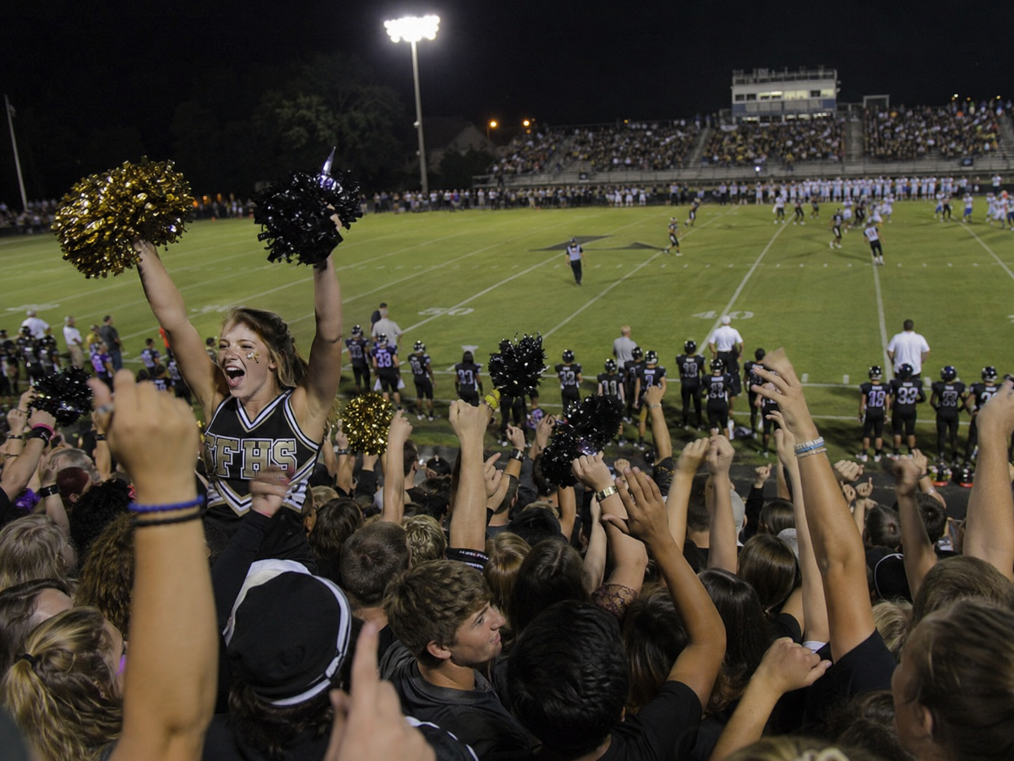 Cheerleader celebrates with gold pom-poms as excited fans rush the field after a victory at a night football game.