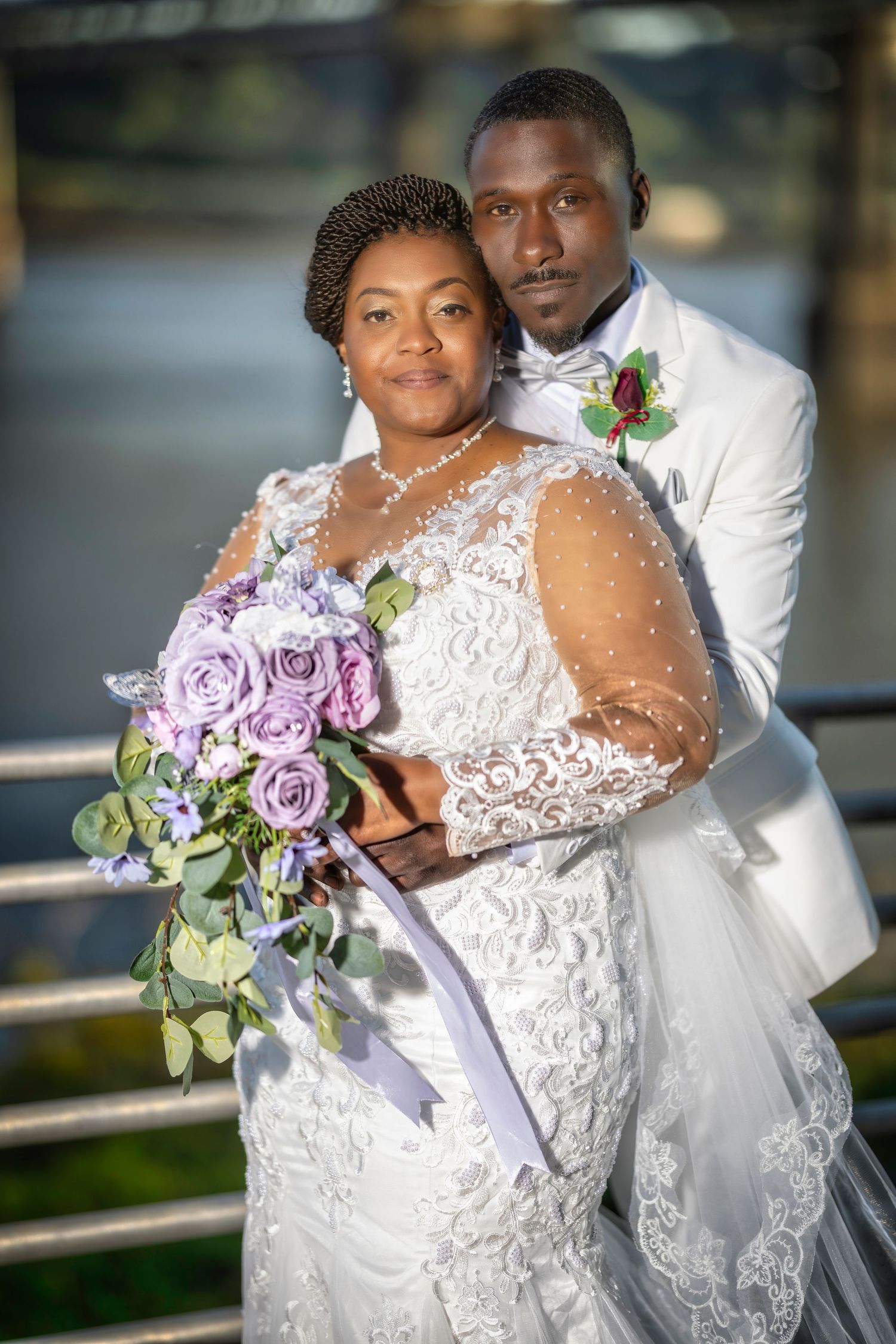 Elegant Black couple in wedding attire, bride in lace gown holding purple roses bouquet, groom in white suit embracing her.