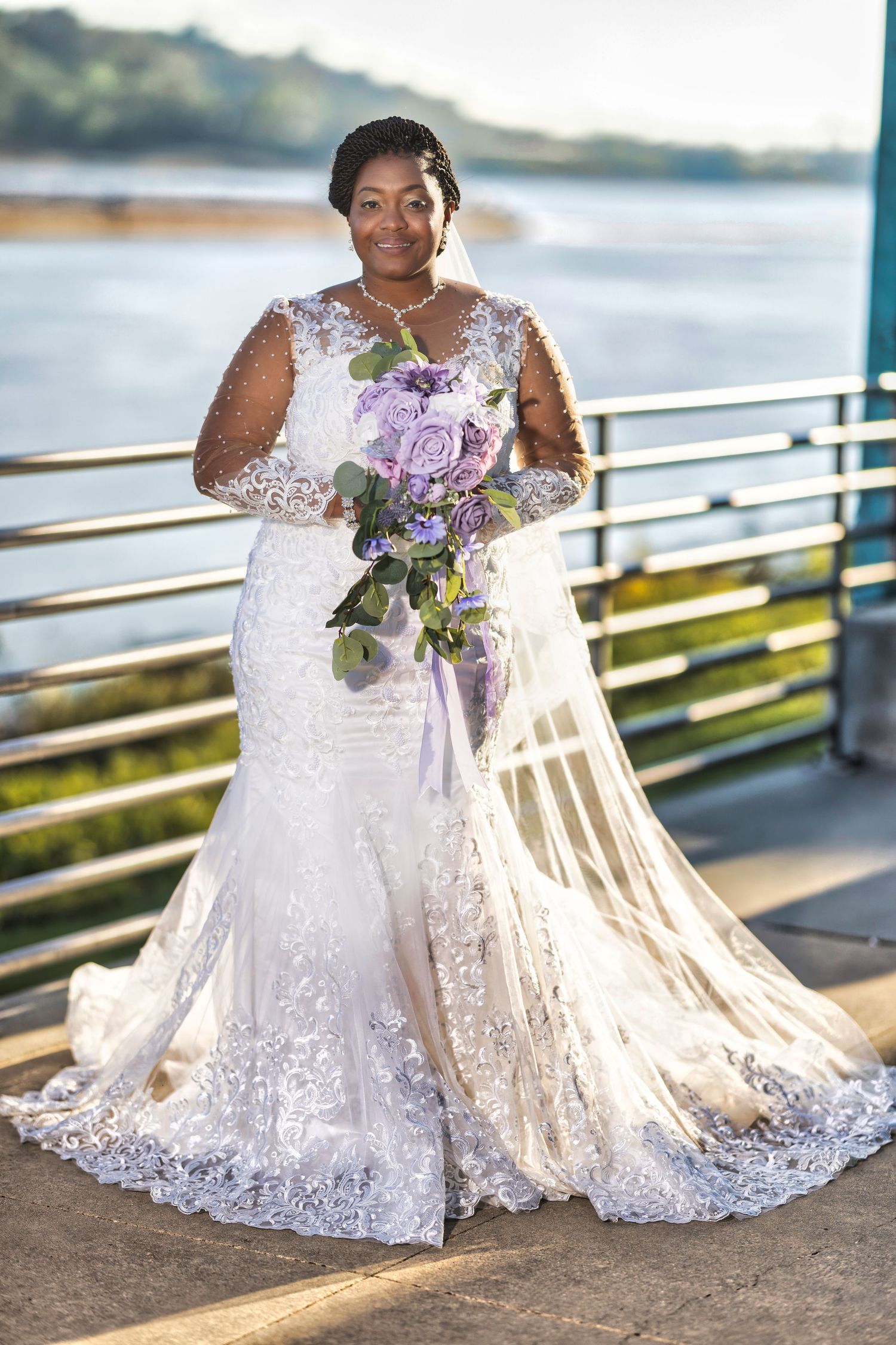 Bride in lace mermaid wedding gown holding purple floral bouquet, posing outdoors near waterfront railing.