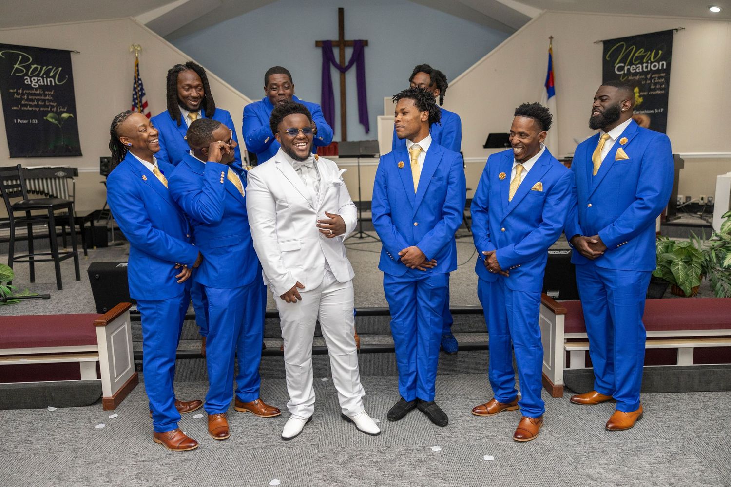 Groom in white suit with groomsmen in royal blue suits and gold ties posing in a church for a wedding photo.