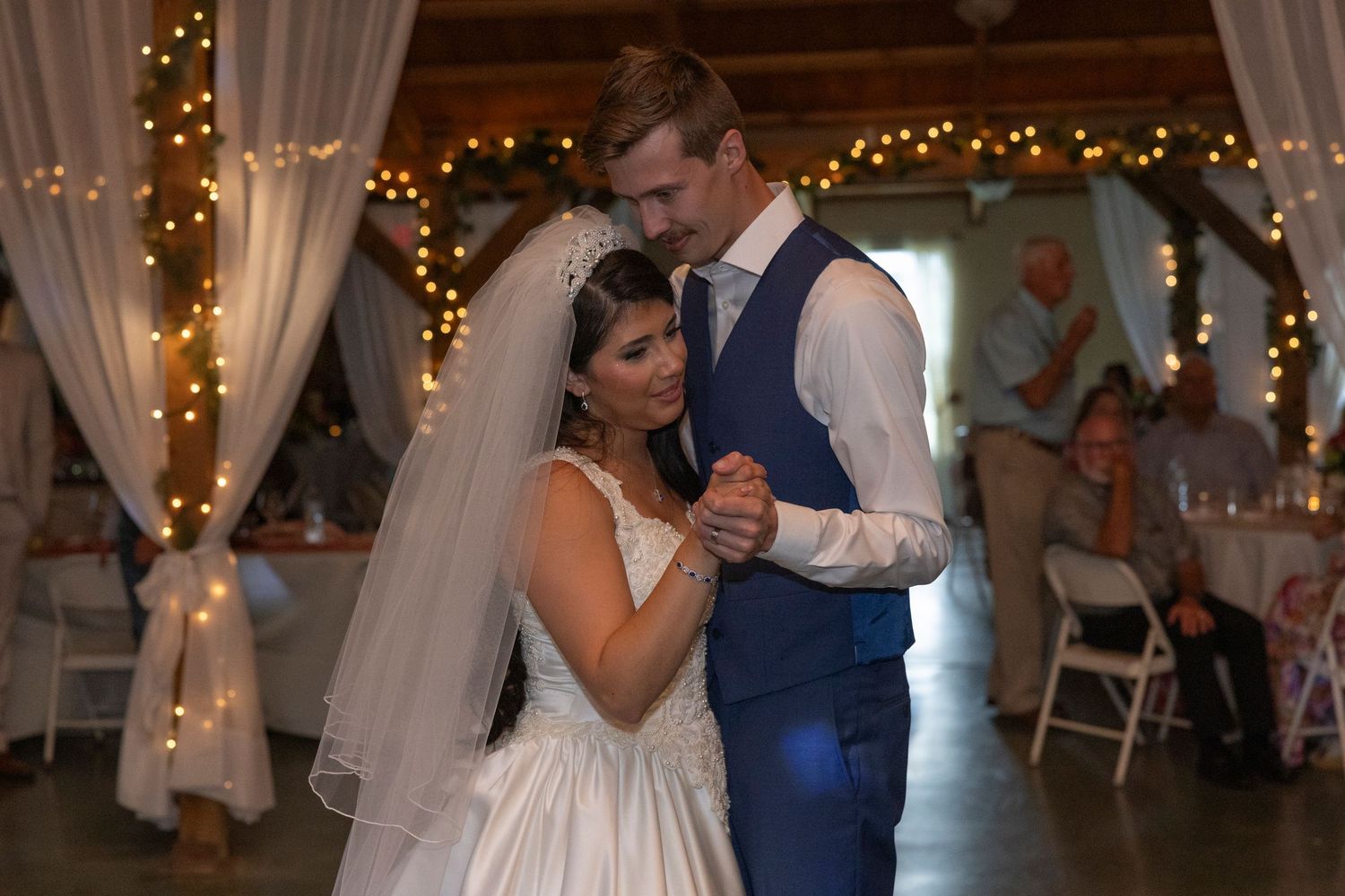 Bride and groom share their first dance at a romantic barn wedding reception with warm string lights and draped fabric.
