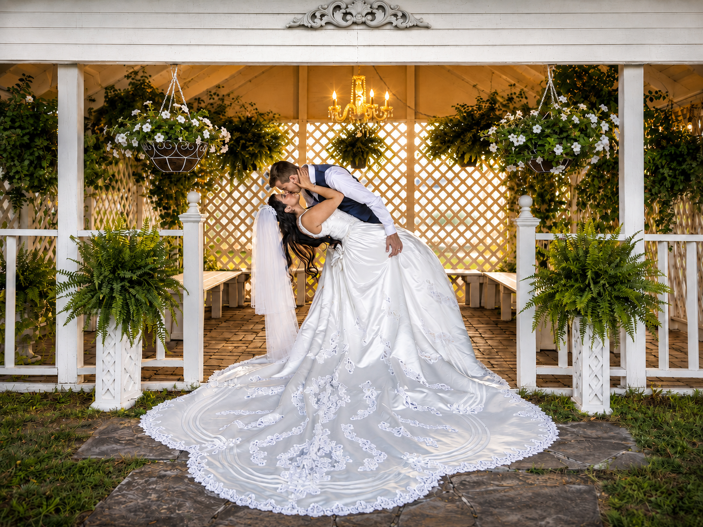 Bride and groom share a romantic dip kiss under a floral gazebo, bride in elegant gown with long lace train.