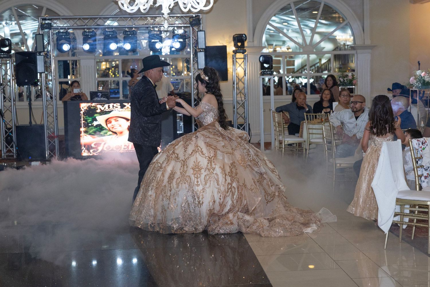 A young woman in a gold ballgown dances with a man at an elegant quinceañera celebration on a misty dance floor.