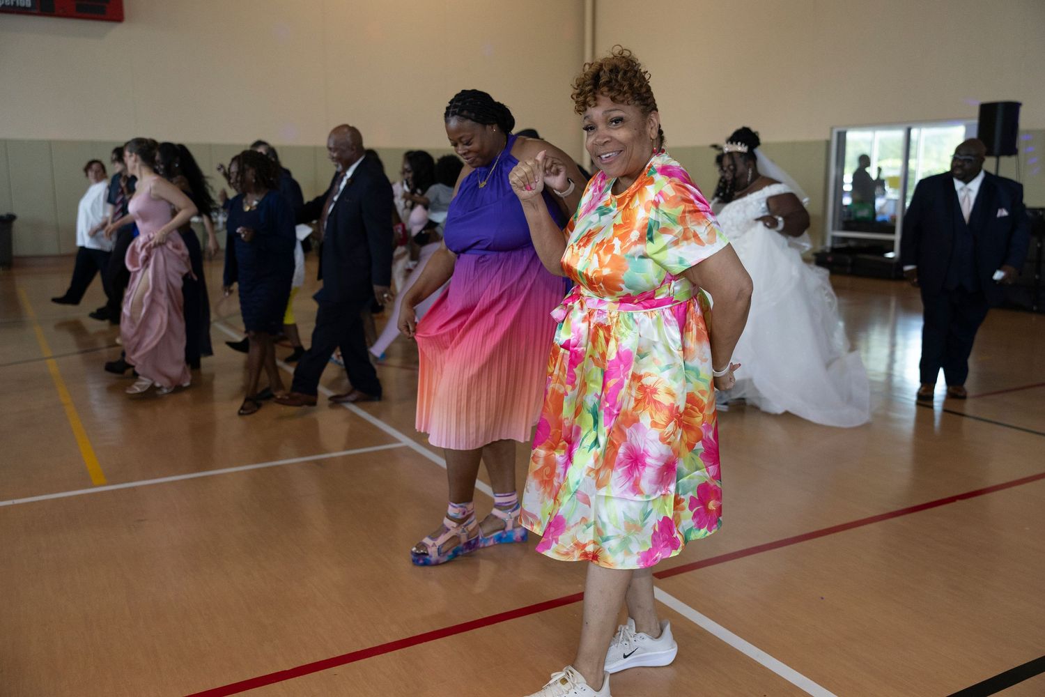 People dancing in colorful dresses at a formal event on a gymnasium floor, with a bride visible in the background.