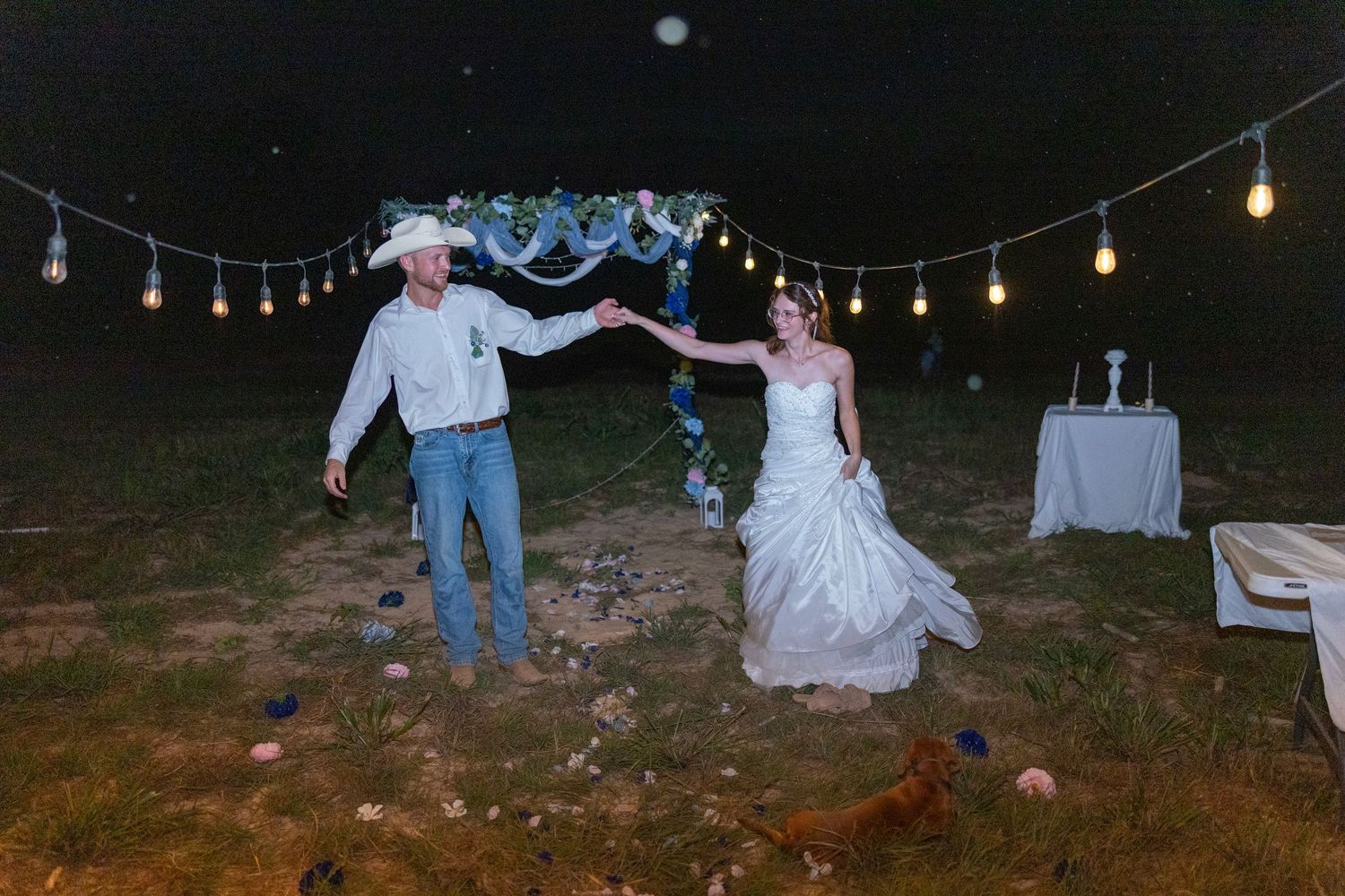 Bride and groom dancing outdoors at night under string lights and a floral arch at a rustic country wedding.