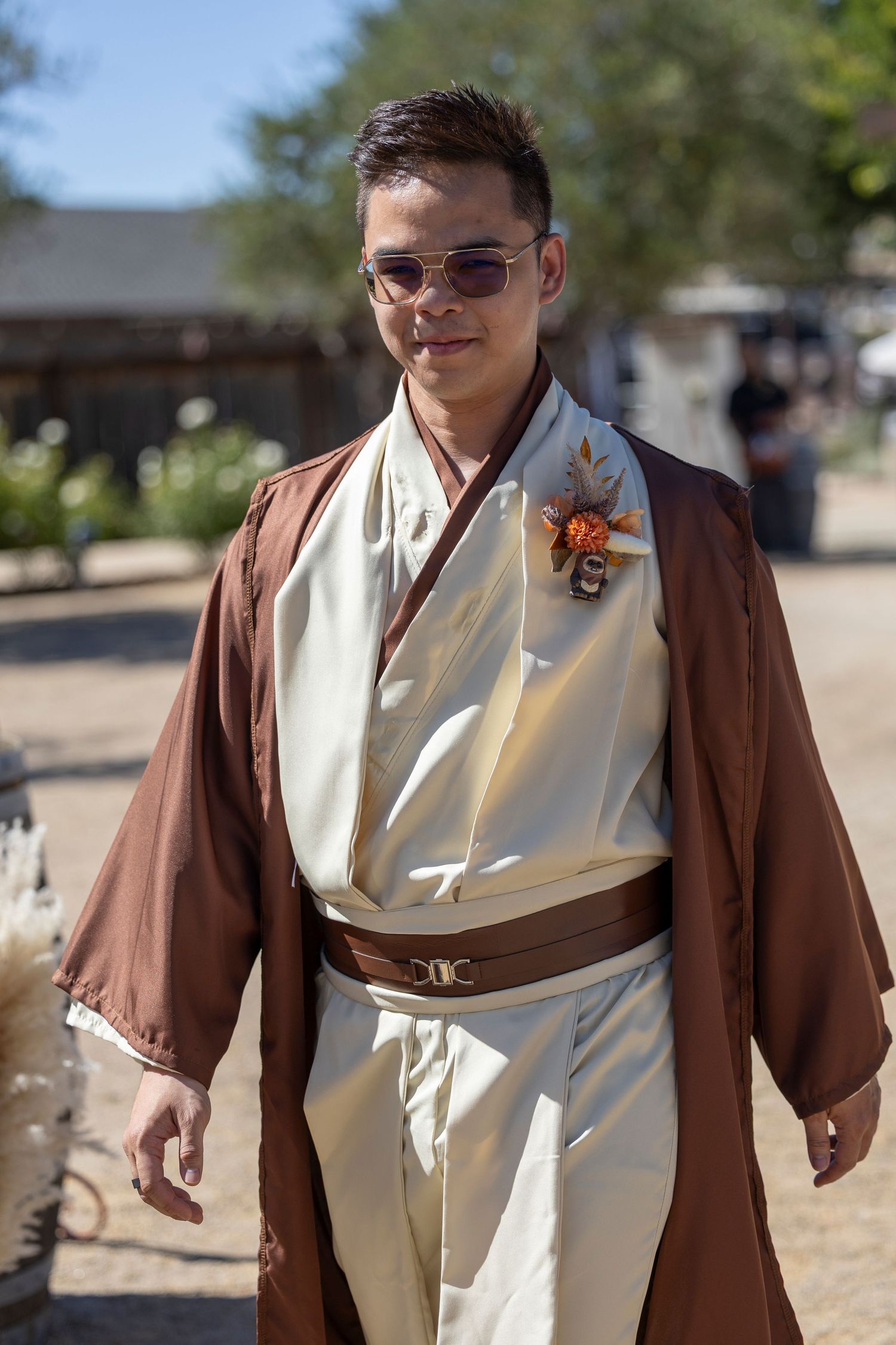 Man wearing a Jedi-inspired wedding outfit with brown robe, cream tunic, belt, and floral boutonniere outdoors.