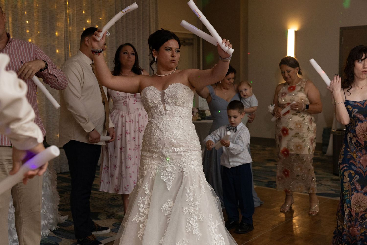 Bride in lace wedding gown dancing with guests holding glow sticks at a festive wedding reception celebration.