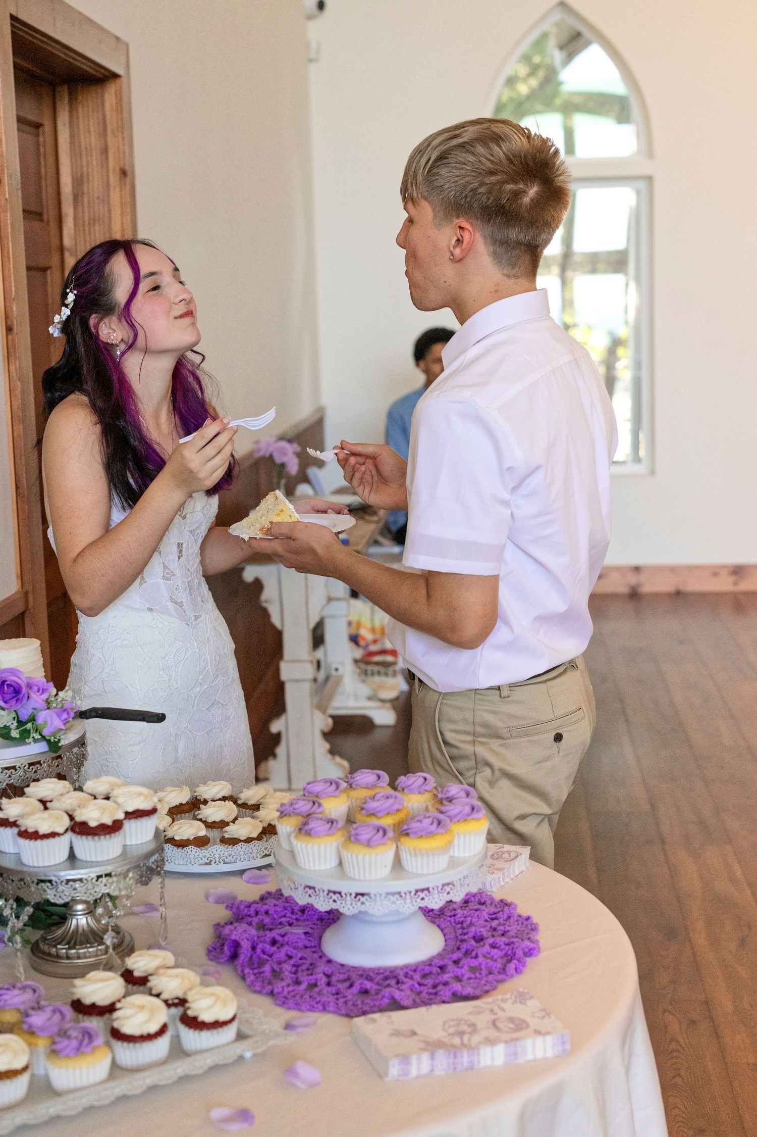 Bride and groom sharing wedding cake at dessert table with purple cupcakes during reception celebration.