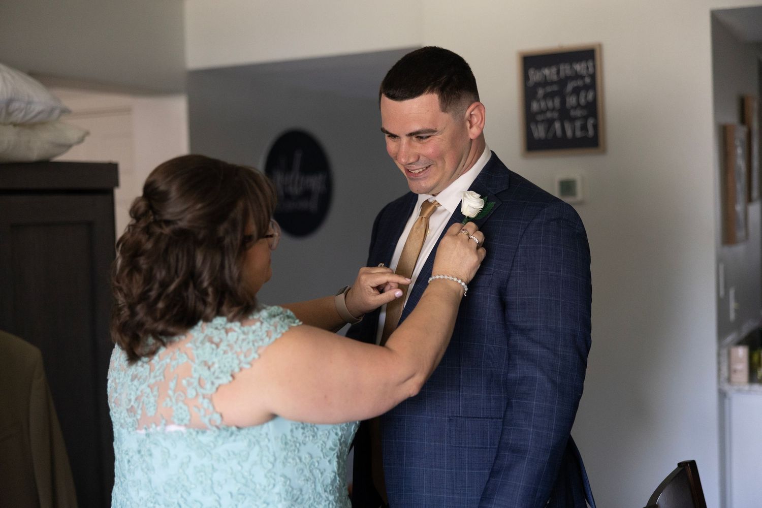 A woman in a blue lace dress pins a boutonniere on a groom in a navy suit with a tan tie.