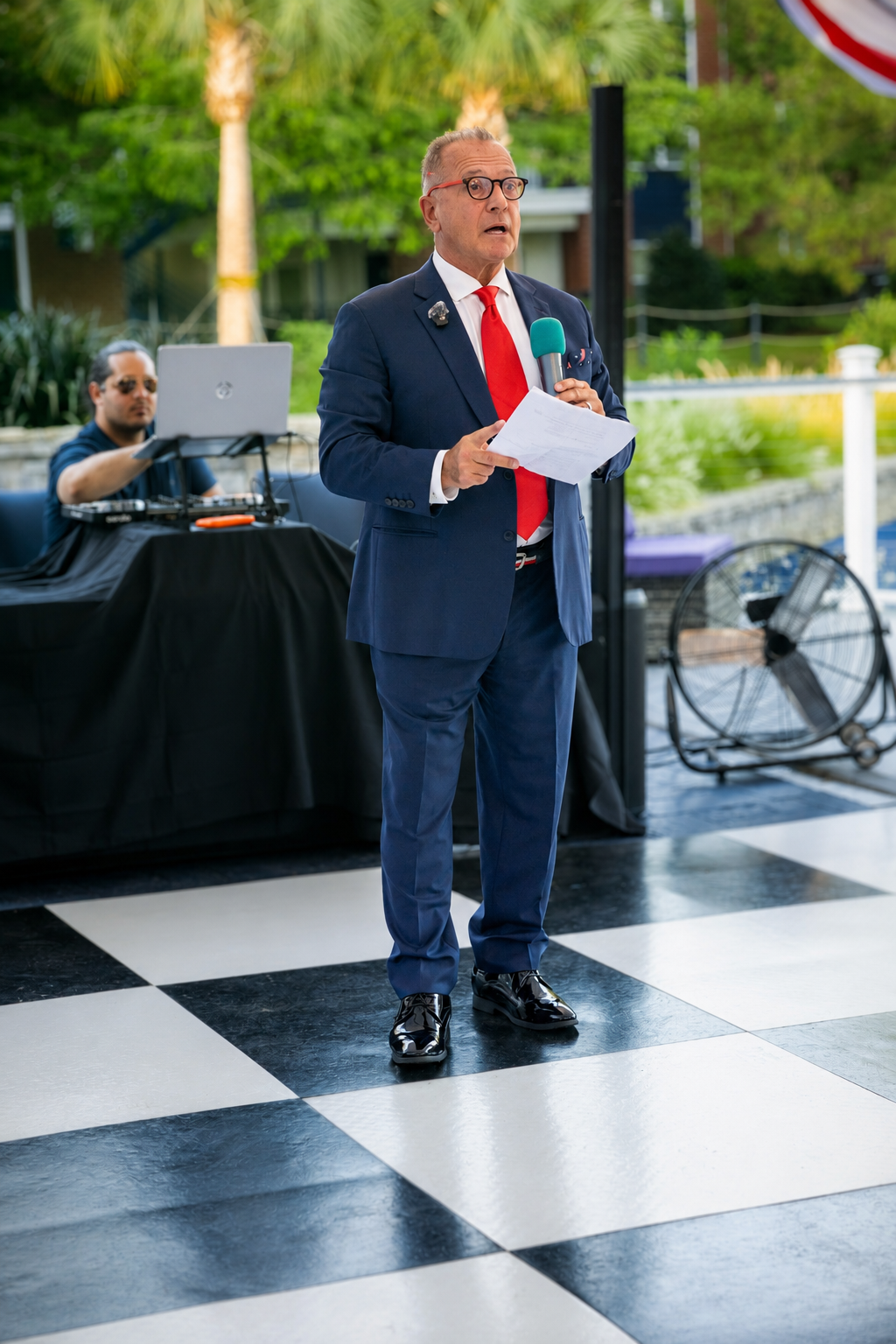 Man in navy suit and red tie speaks at outdoor event on checkered dance floor holding microphone and papers.