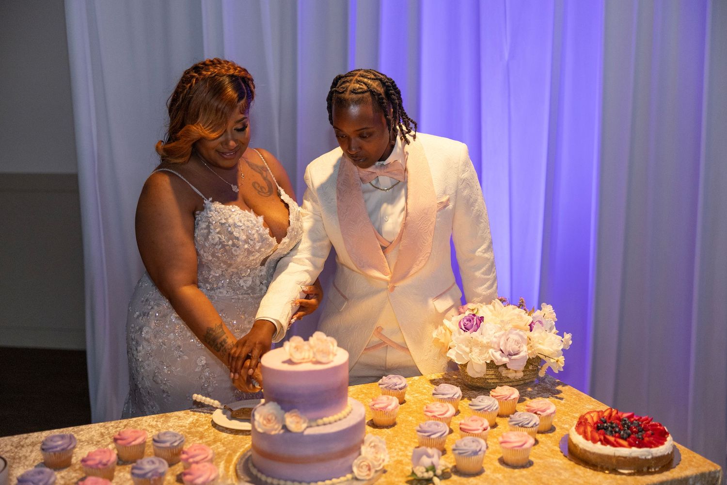 Bride and groom cutting wedding cake together at reception table with cupcakes and floral arrangements.