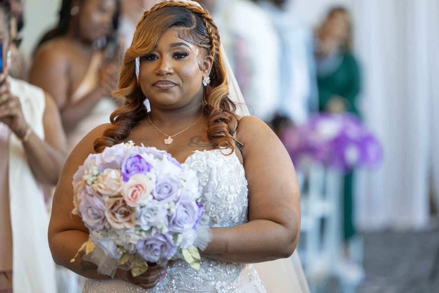 Bride in beaded gown holding purple and pink bouquet walks down aisle at wedding ceremony.