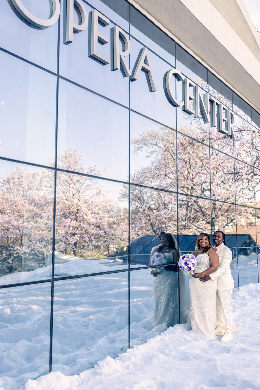 Bride and groom pose in snow outside Opera Center with cherry blossoms reflected in glass windows.