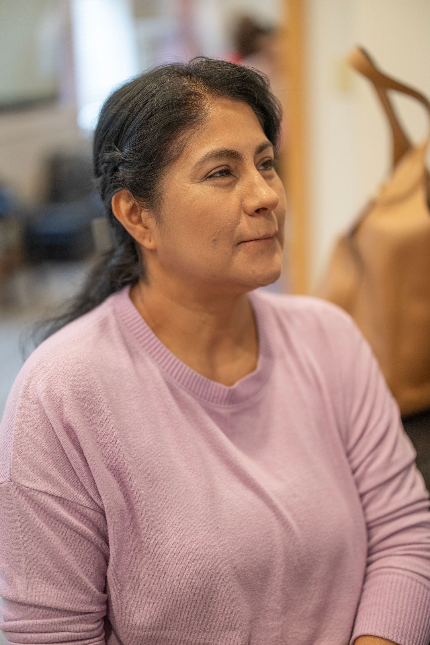 Middle-aged woman in pink sweater sitting indoors, looking thoughtfully to the side in a casual setting.