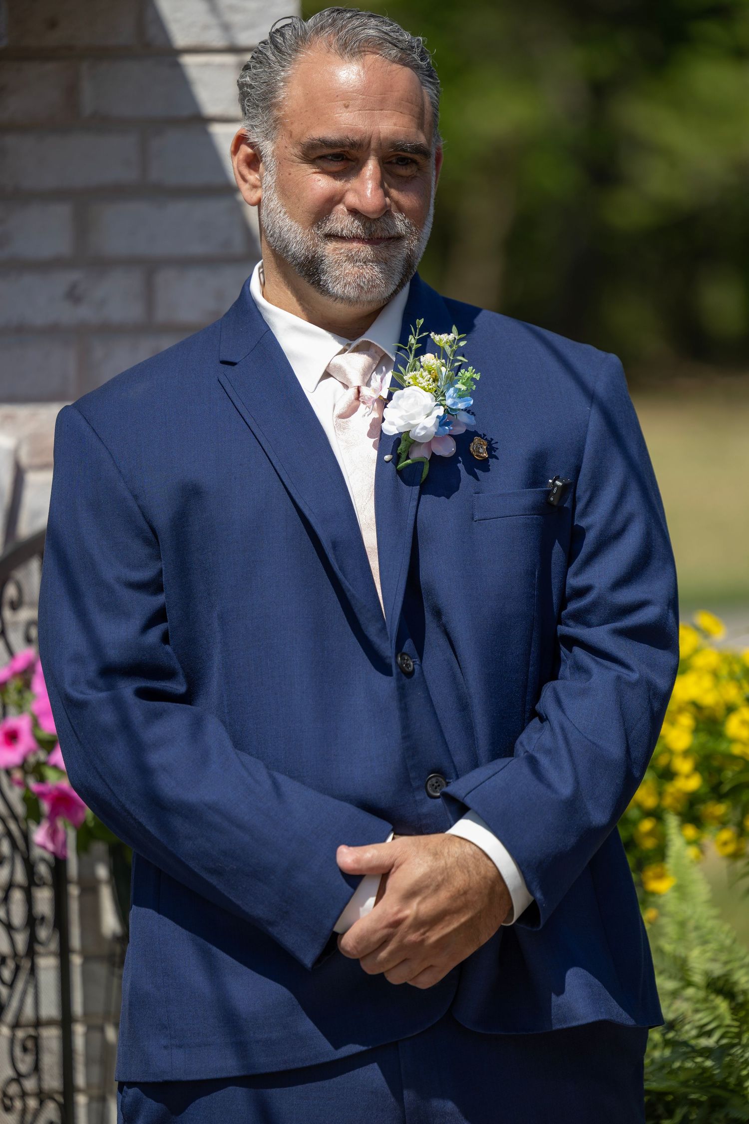 Man in navy blue suit with floral boutonniere smiling outdoors at a wedding ceremony.