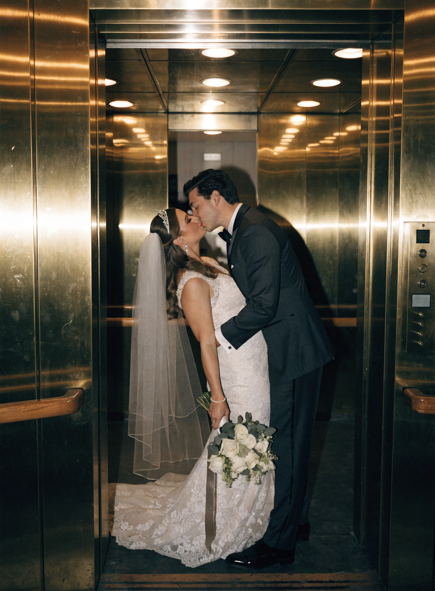 Bride and groom share a romantic kiss in a gold elevator, bride holding white bouquet and wearing lace gown with veil.