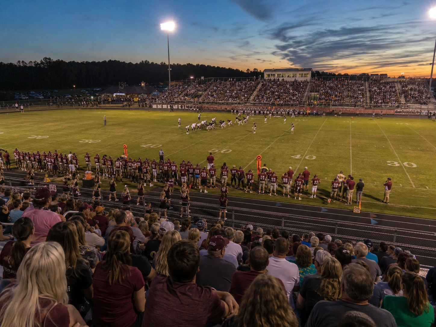 Packed high school football stadium at sunset with players on the field and crowds filling the bleachers.