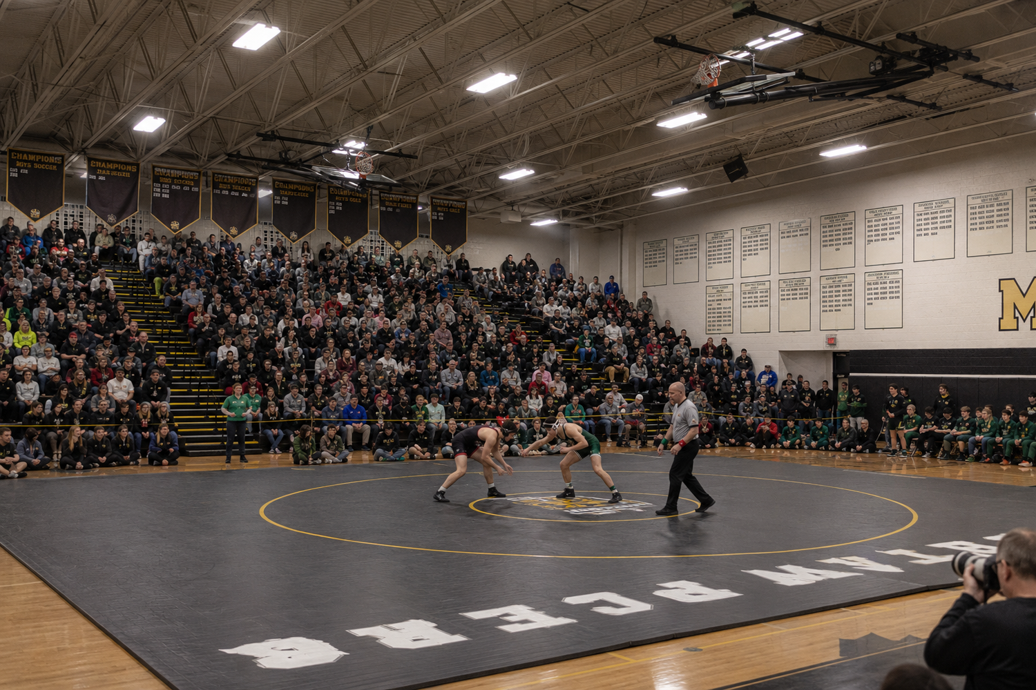 Wide view of high school wrestling match on gym floor with large crowd filling bleachers in decorated gymnasium.