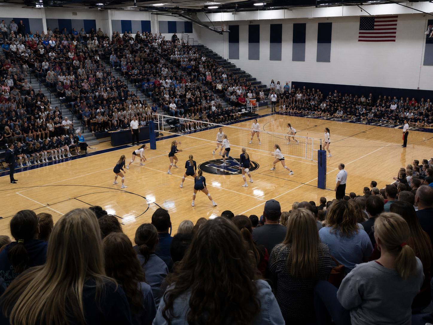 Packed gymnasium crowd watches intense high school volleyball match on hardwood court.