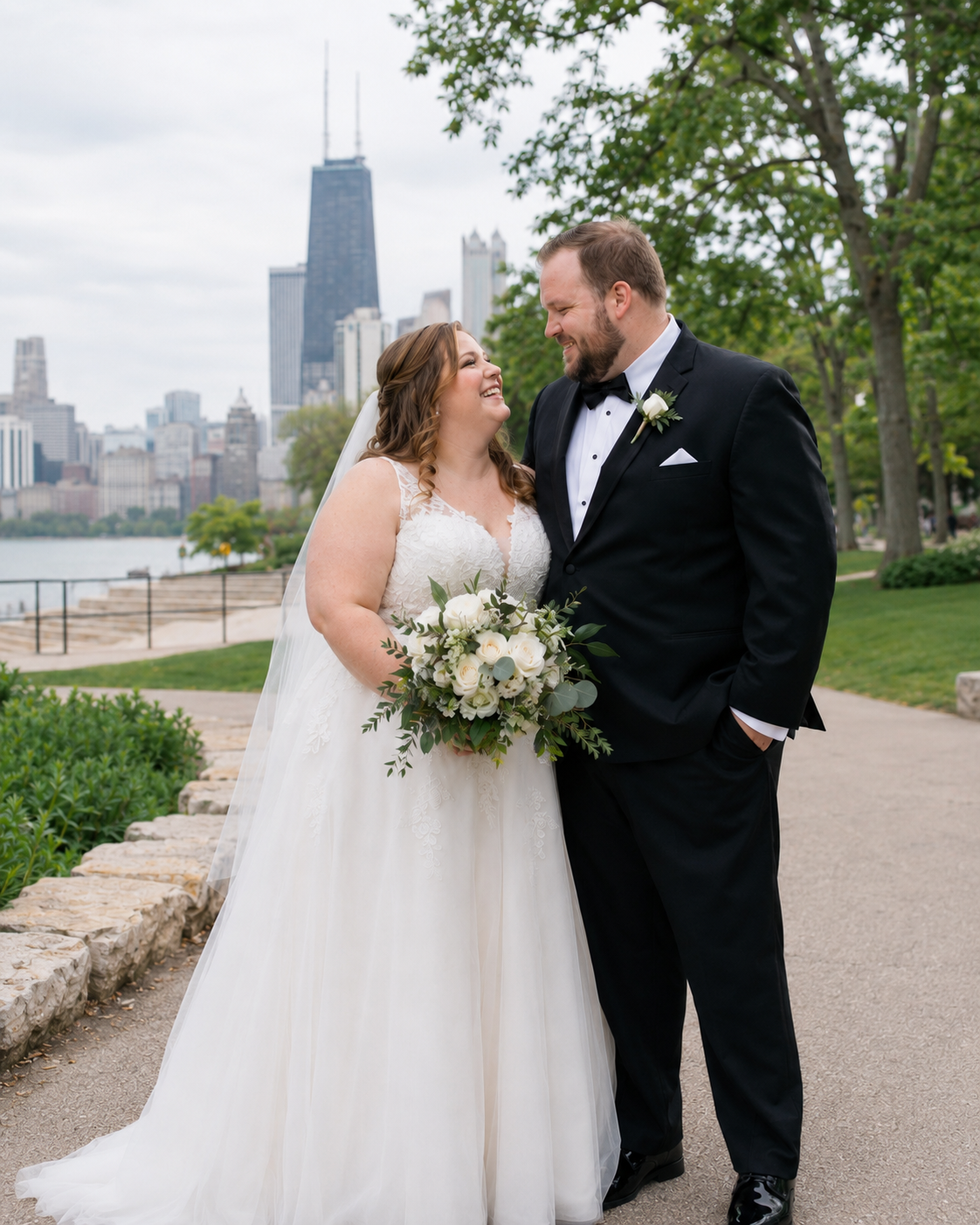 A bride and groom smile at each other in front of the Chicago skyline, she holds a white bouquet.