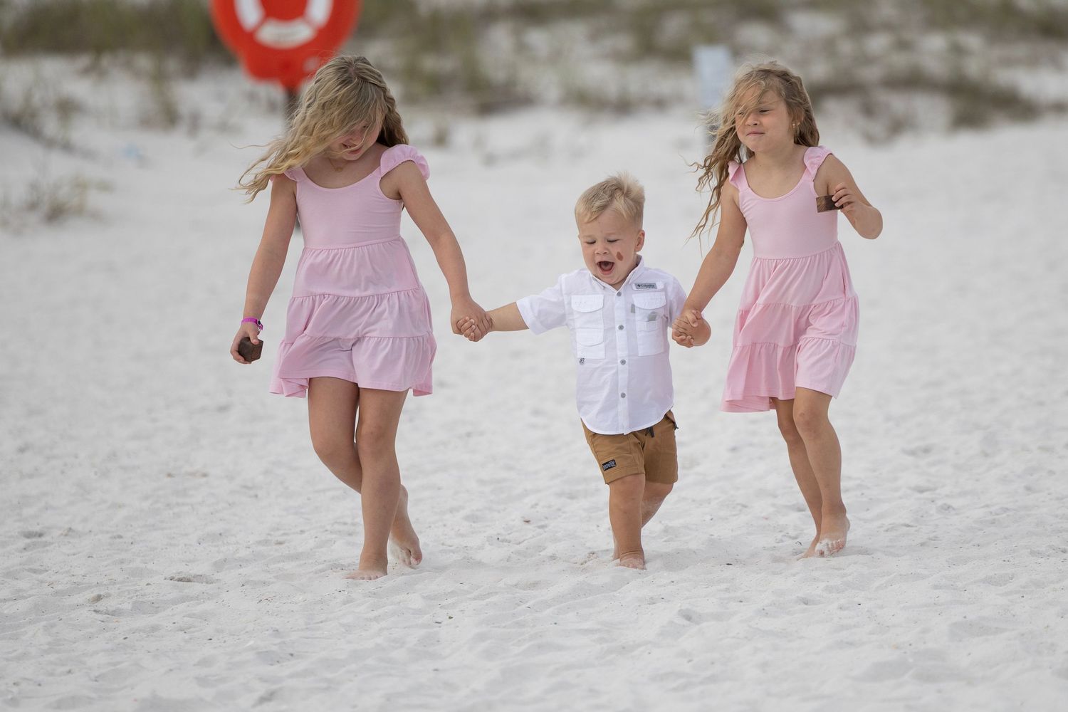 Three young children in pink outfits joyfully running and holding hands on a sandy beach.