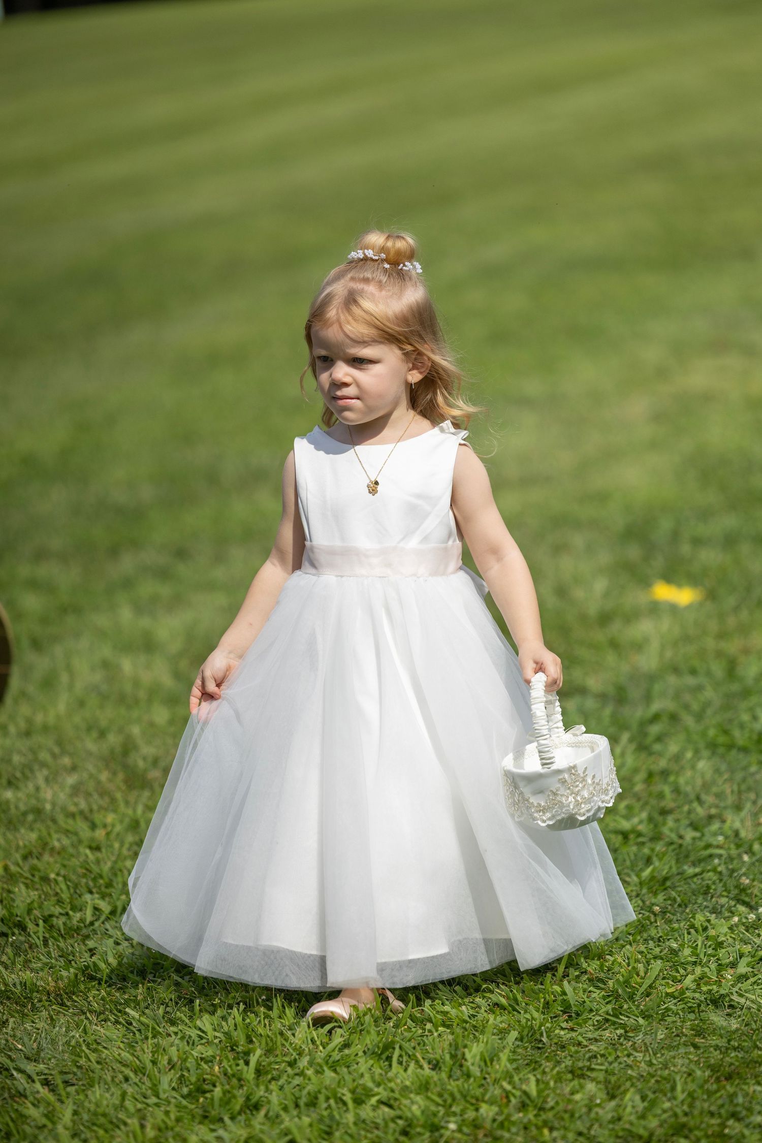 A small child in a white dress and flower crown plays on a grassy lawn during a sunny day.