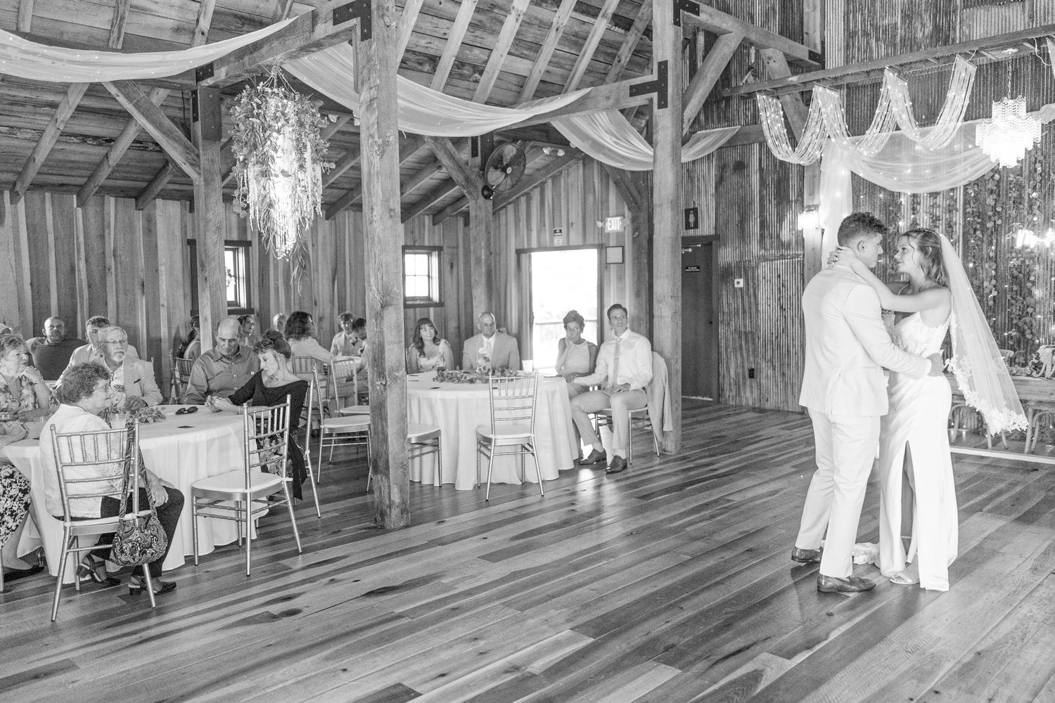 A couple shares a romantic dance on a rustic wooden floor at a barn wedding reception with guests seated at tables.
