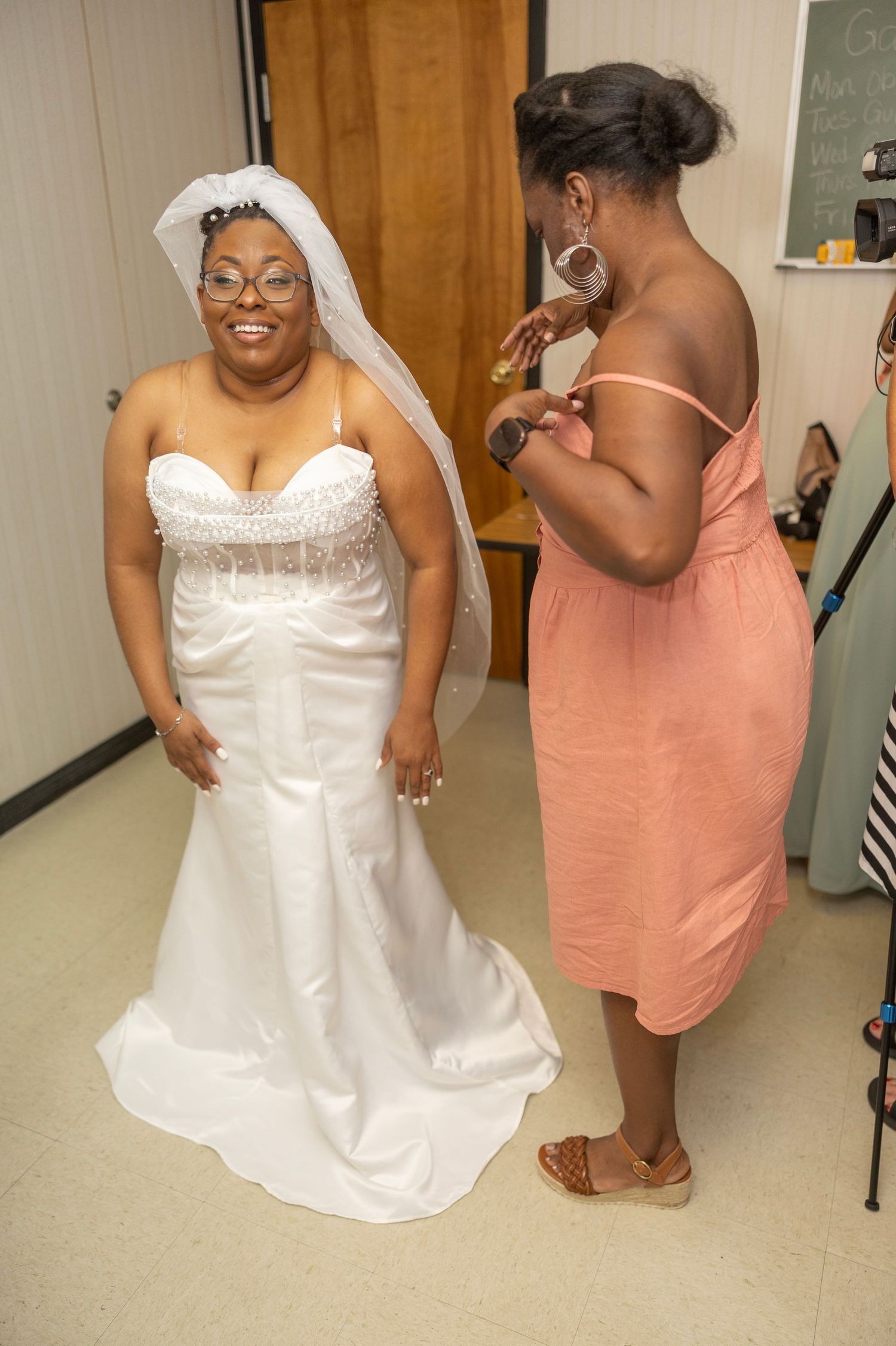 A bride in a white strapless wedding gown poses with a bridesmaid wearing a peach-colored dress in a hallway.