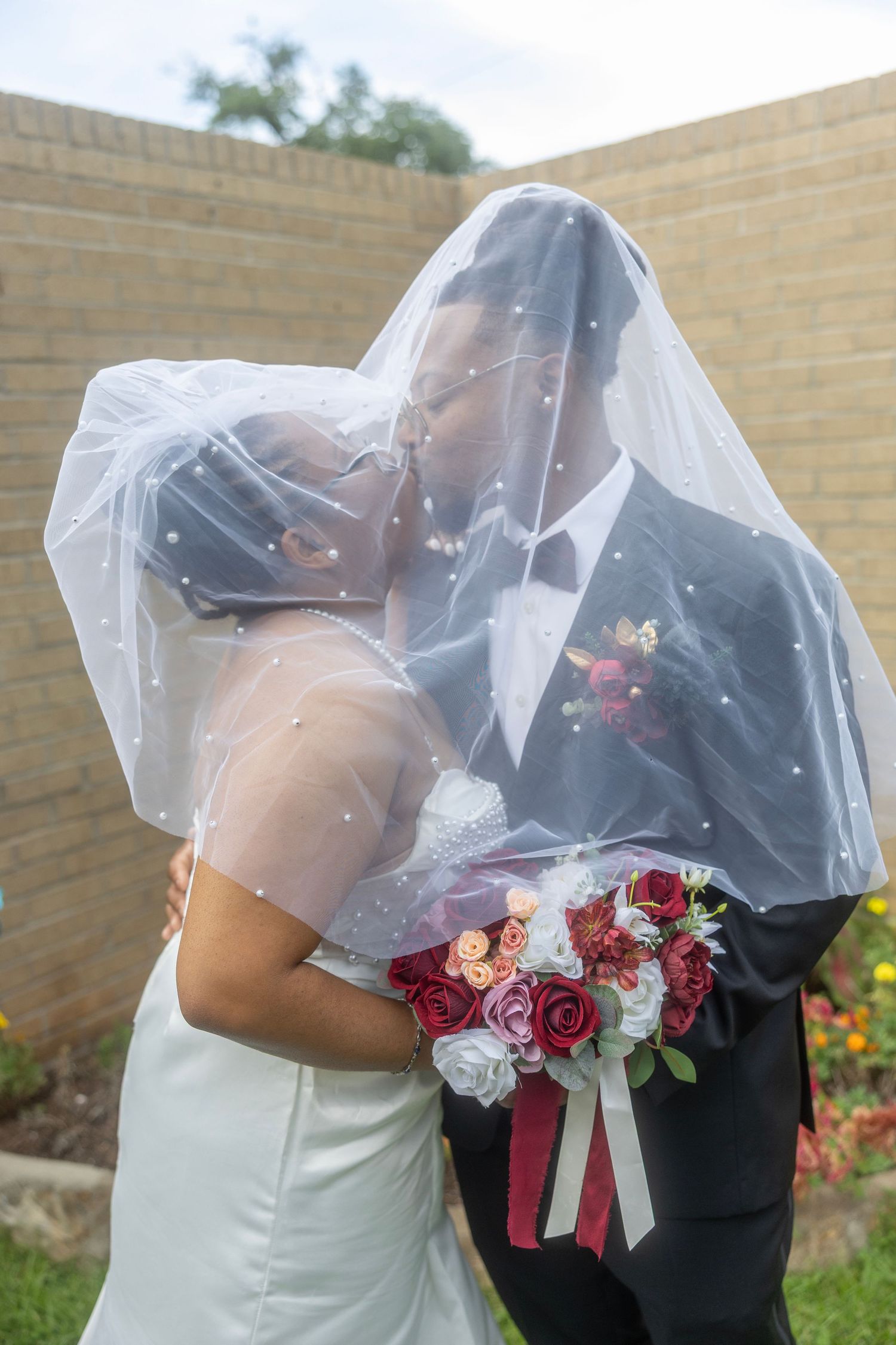 Newlyweds share a romantic moment under a sheer veil while holding a vibrant bouquet of red and pink roses.