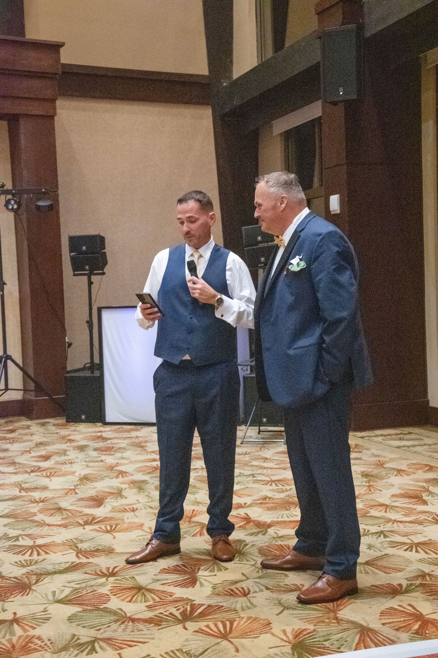 Four groomsmen in matching navy suits and brown shoes stand on a patterned carpet floor while looking at their phones.
