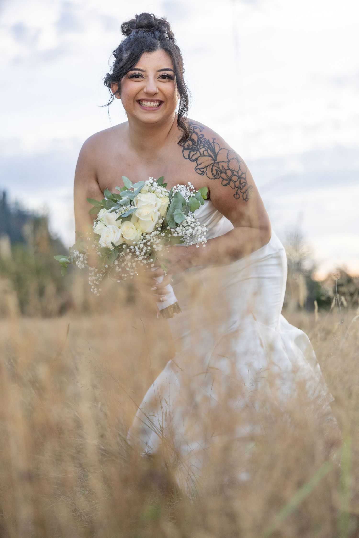A bride in a white strapless wedding dress smiles joyfully while holding a bouquet in a sunlit field of tall grass.