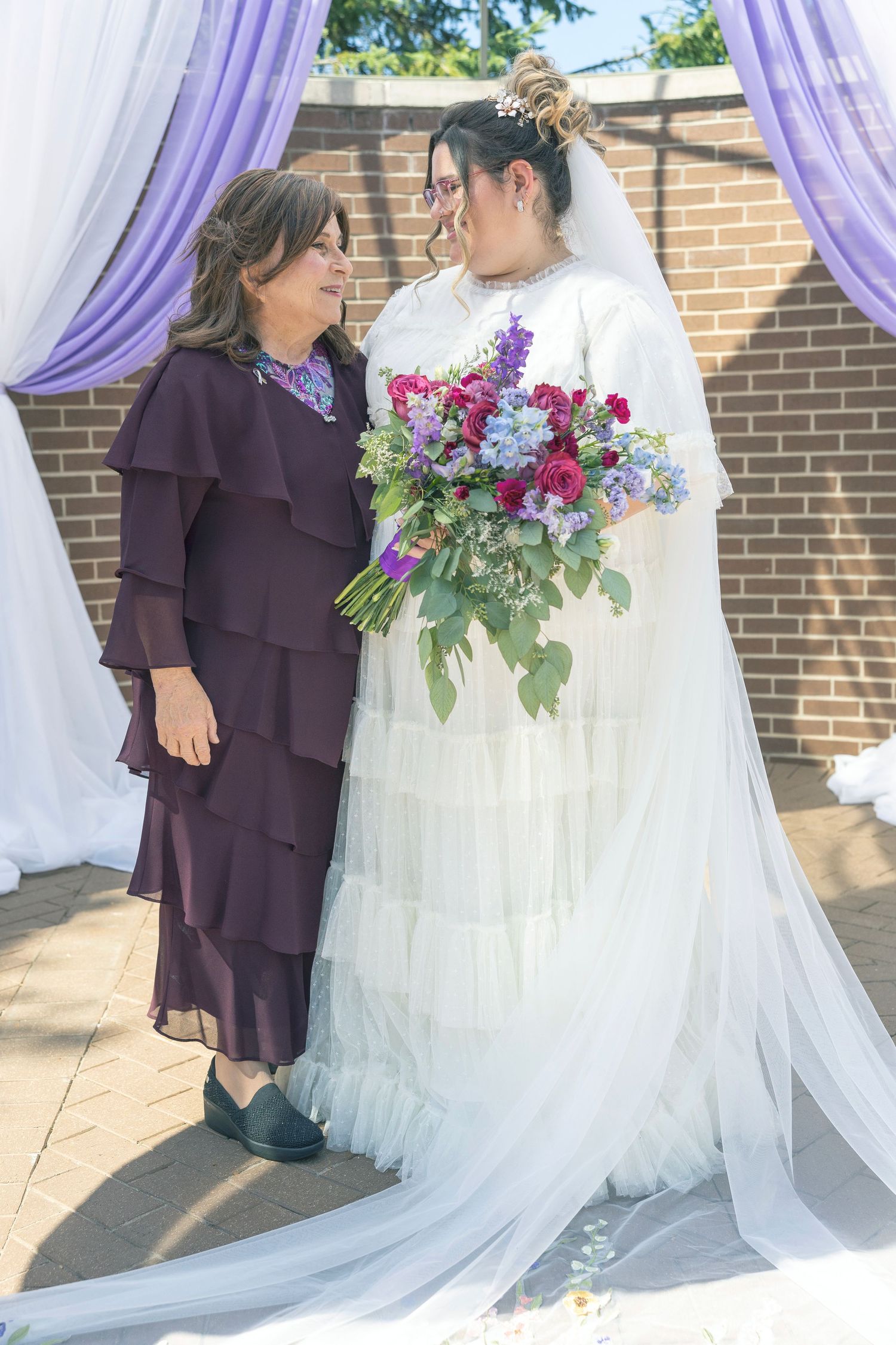 Wedding party members in purple and white dresses pose together with vibrant floral bouquets against a brick wall.