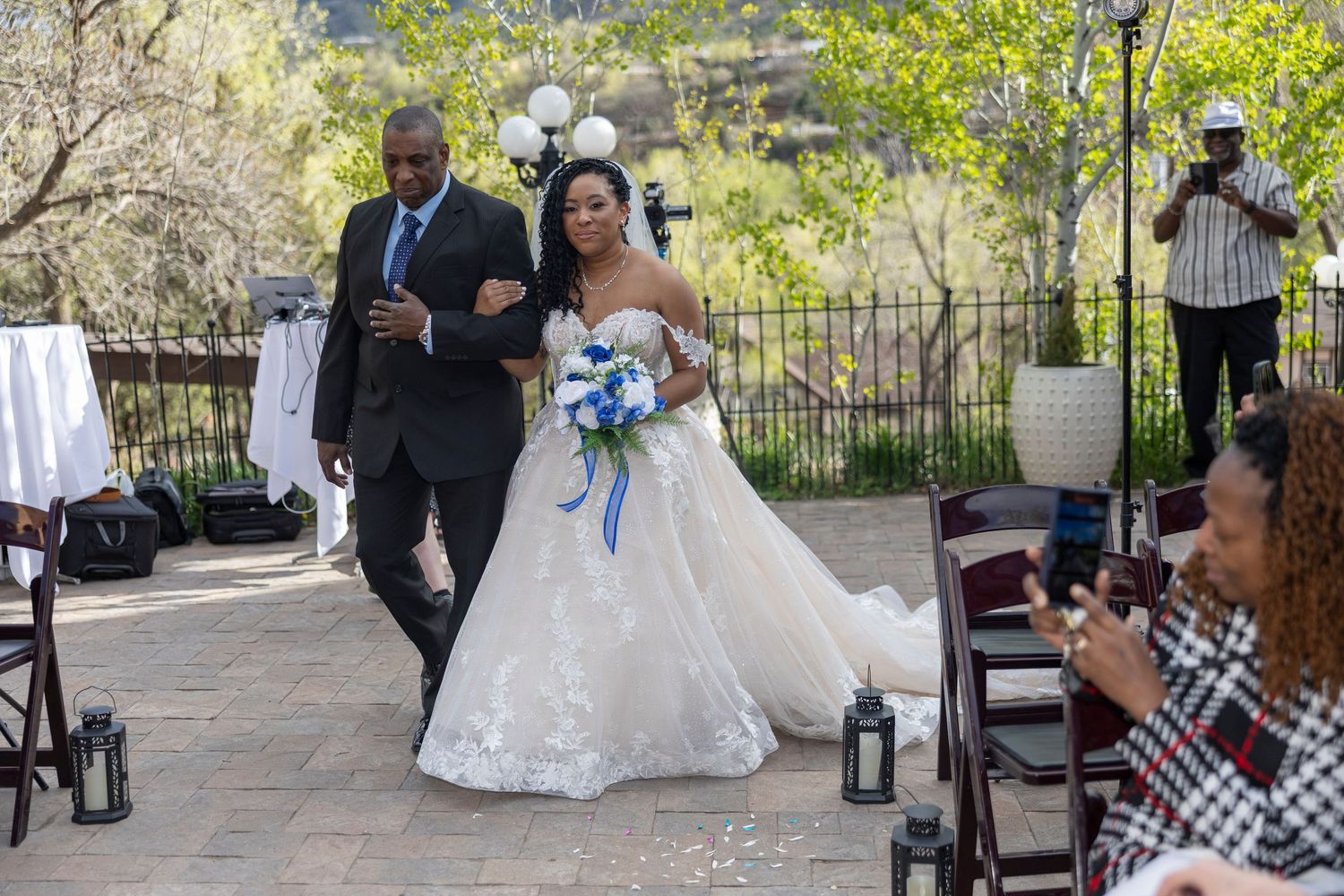 Wedding ceremony taking place in an outdoor garden setting with guests seated on white chairs and floral decorations.