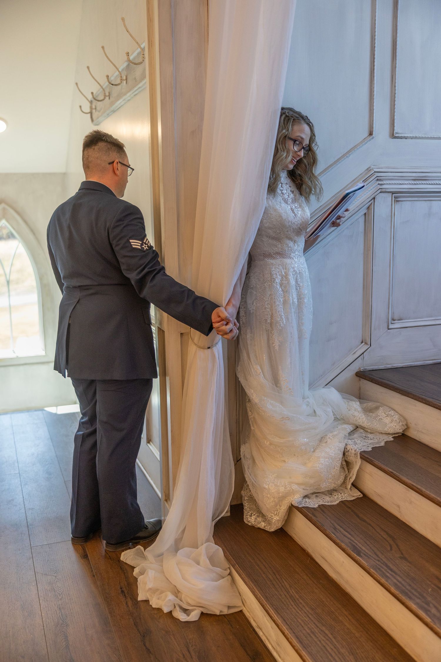 A bride in a flowing white dress ascends wooden stairs while holding hands with a groom in a dark suit.