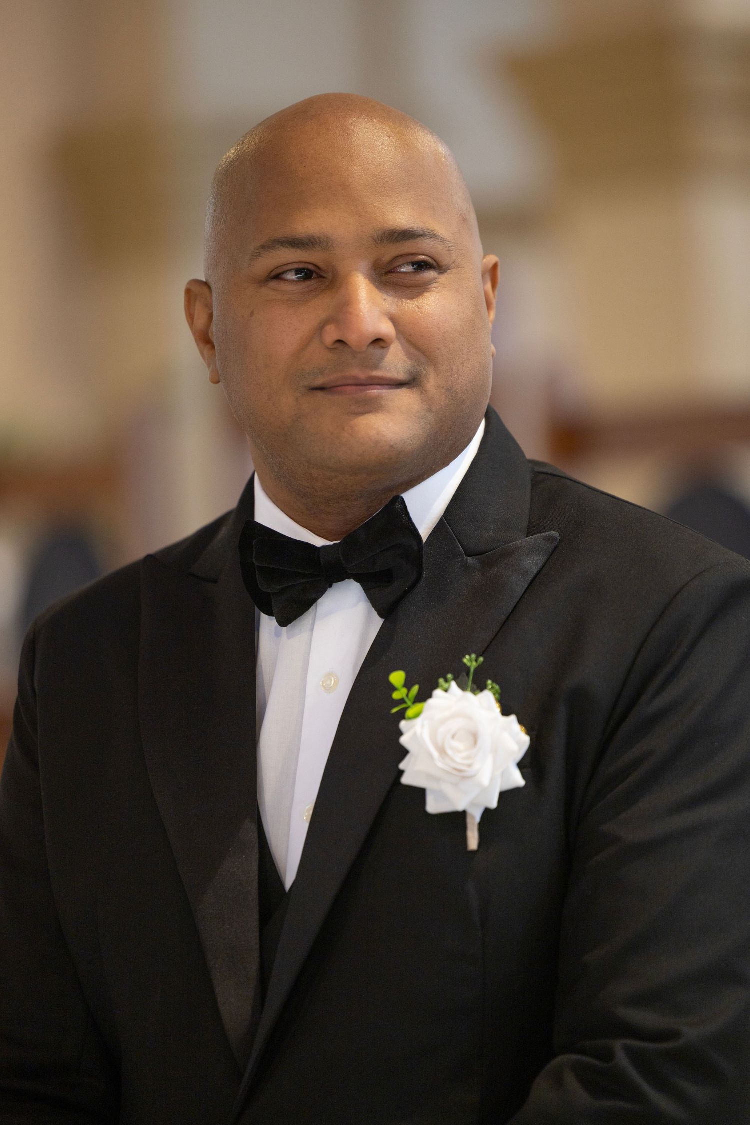 A groomsman wearing a black tuxedo with a white boutonniere smiles in a formal wedding portrait.