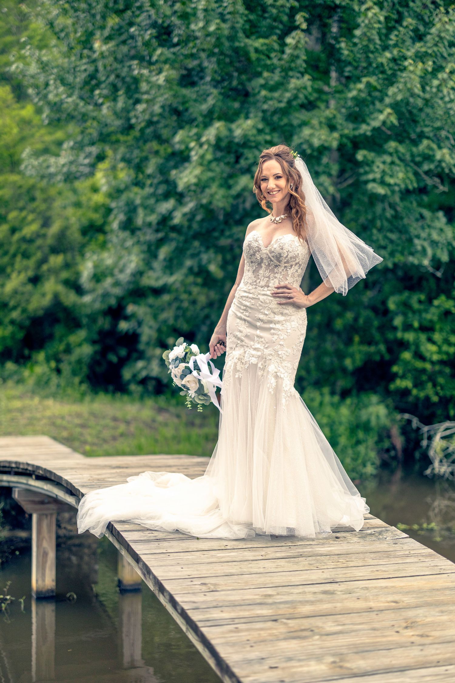 A bride in a flowing white dress poses playfully on a wooden dock by a serene lake surrounded by greenery.