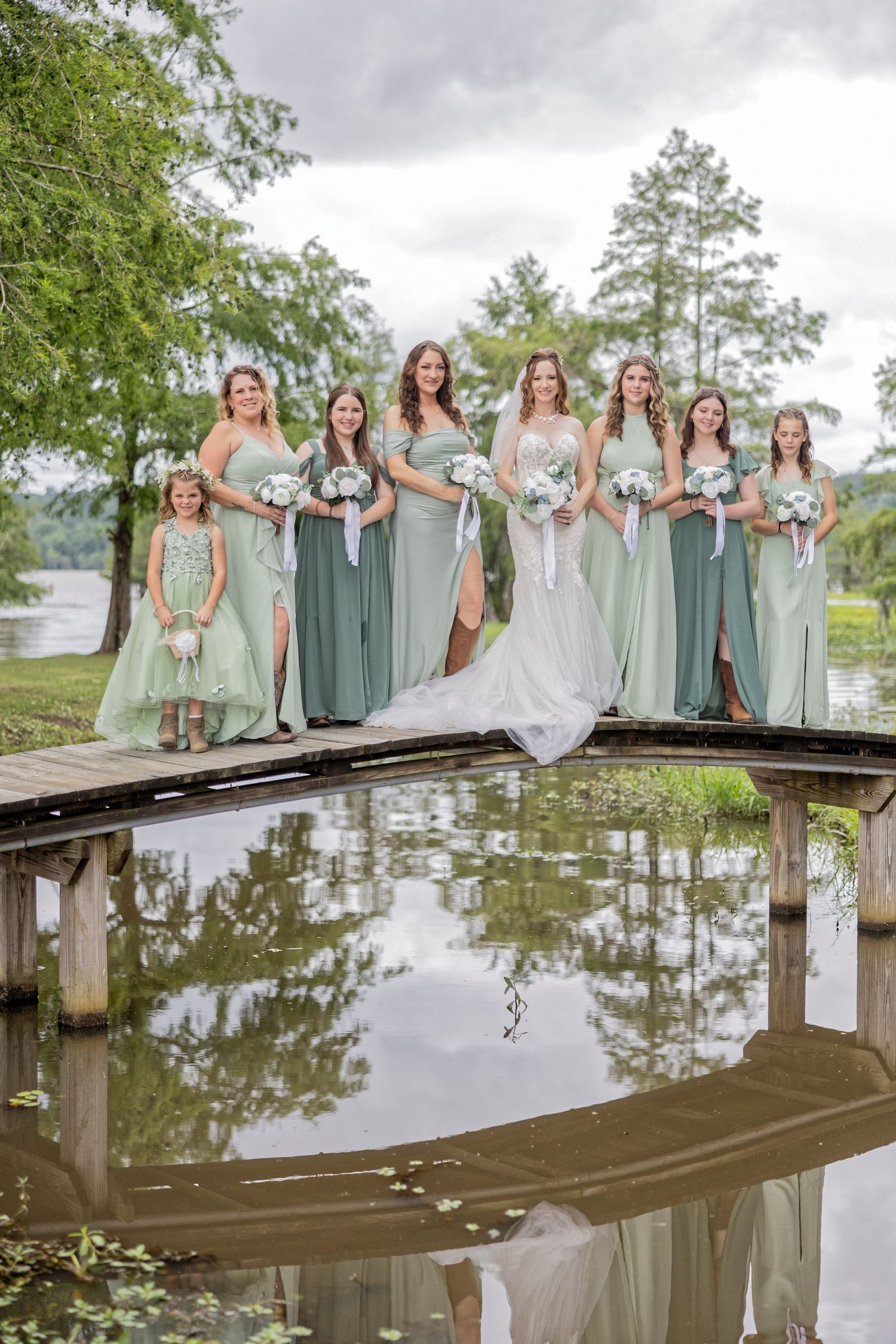 Wedding party in sage green dresses poses together on wooden dock over reflective lake for outdoor bridal photos.