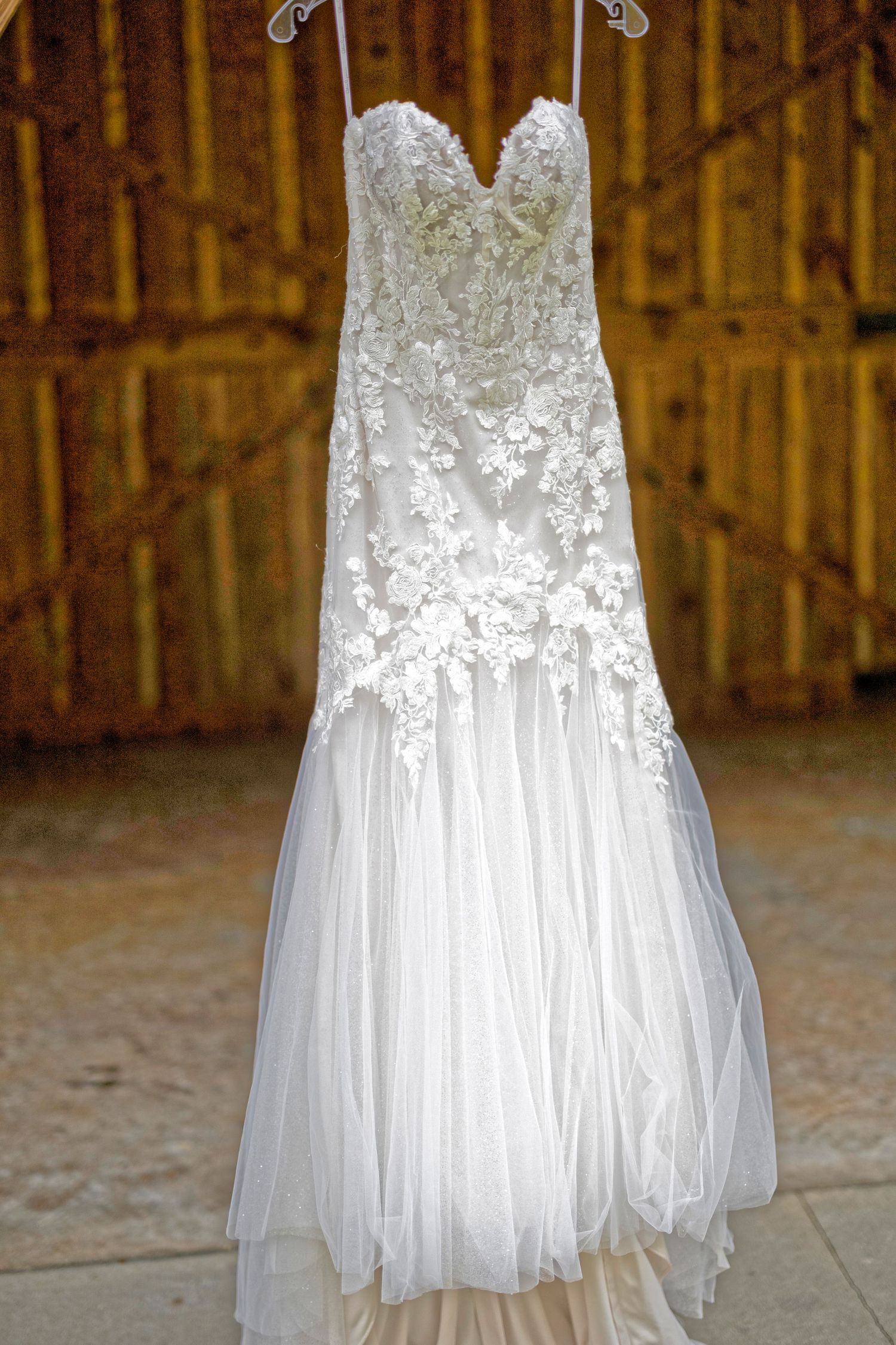 White wedding dress with delicate lace appliques and flowing tulle skirt hangs in rustic barn setting.