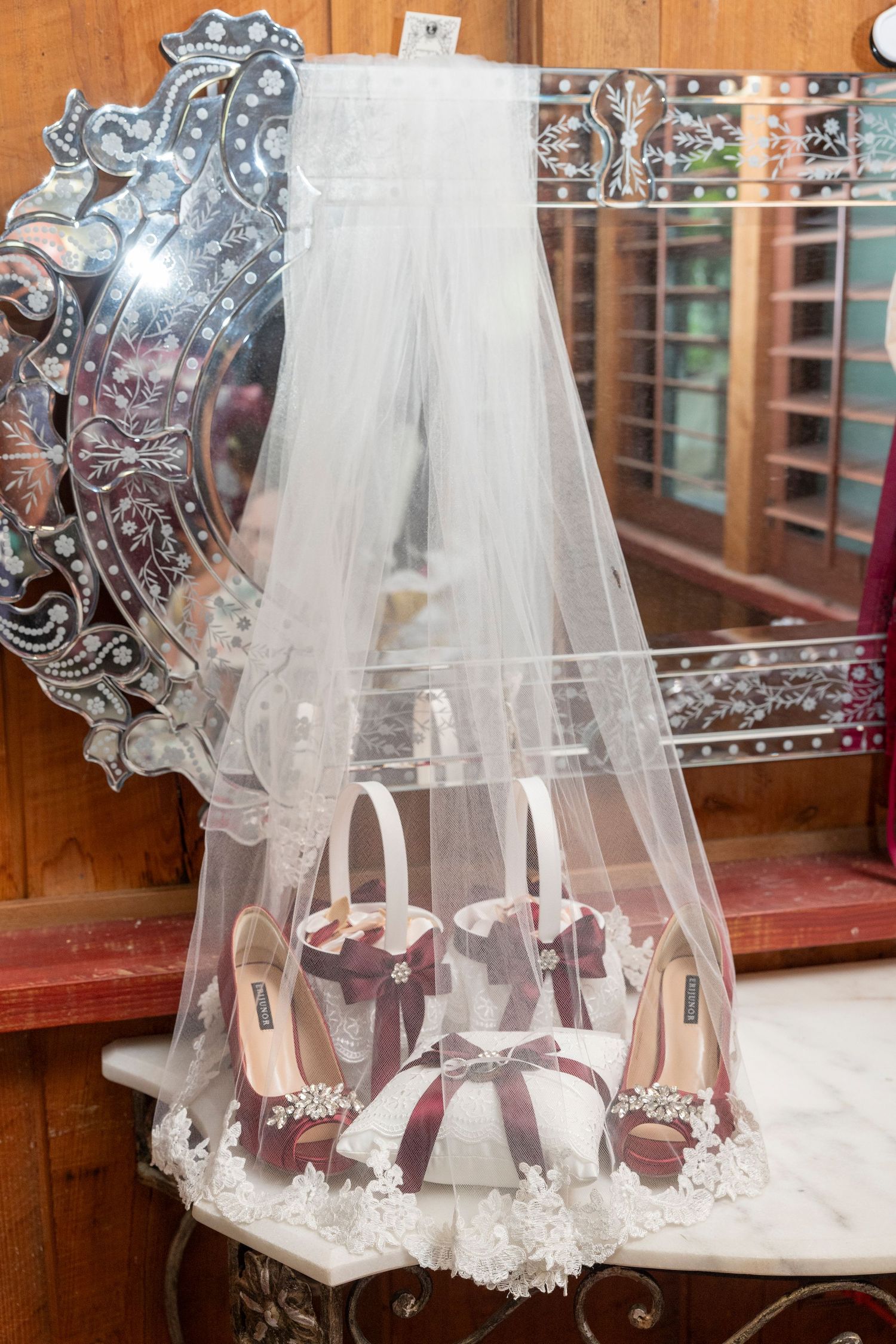 Wedding veil and burgundy high heel bridal shoes displayed on ornate mirrored vanity with decorative details.